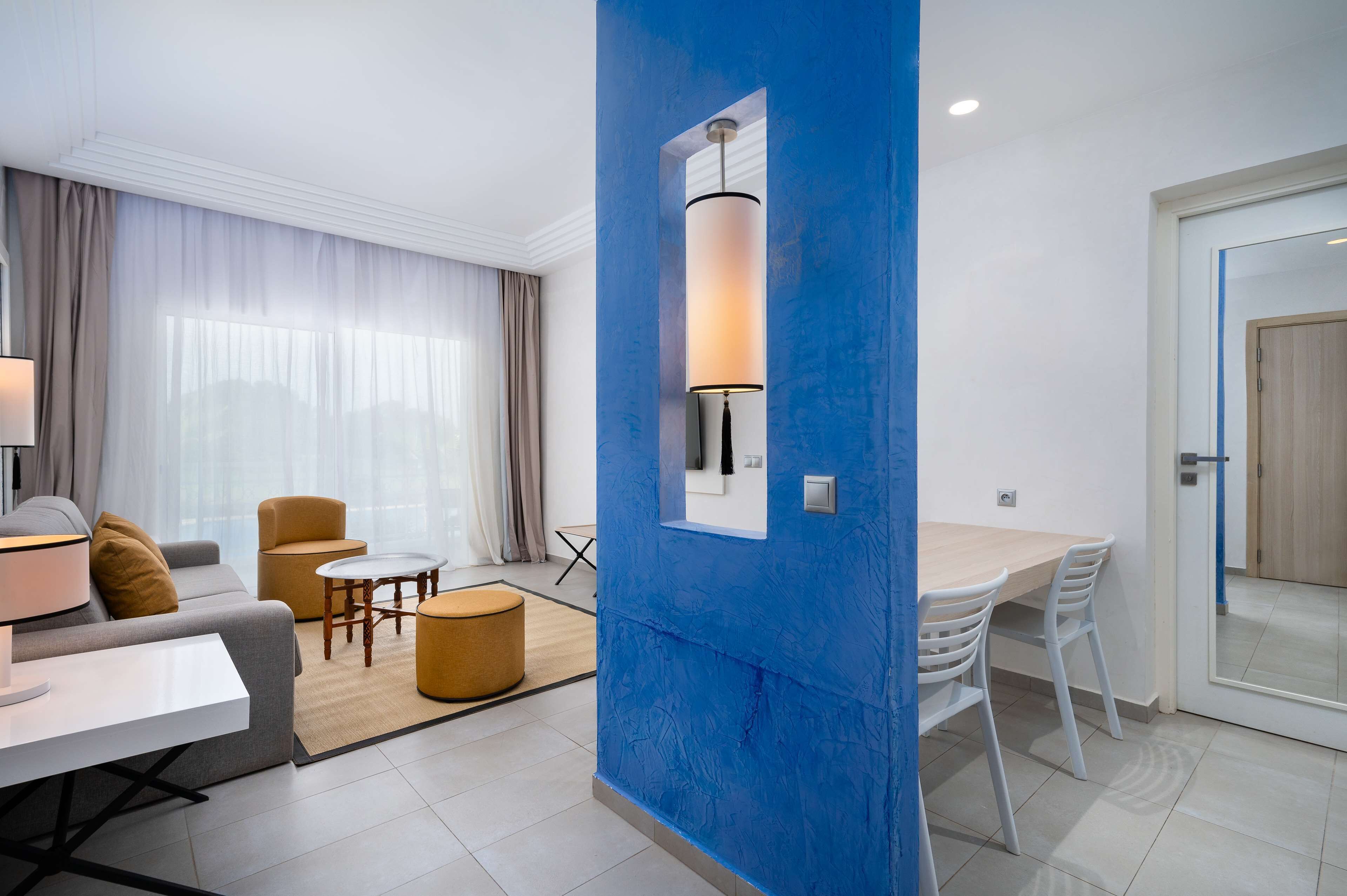 Habitación de hotel moderna con un ventanal grande, una pared de acento azul, zona de comedor y sofá con lámpara.