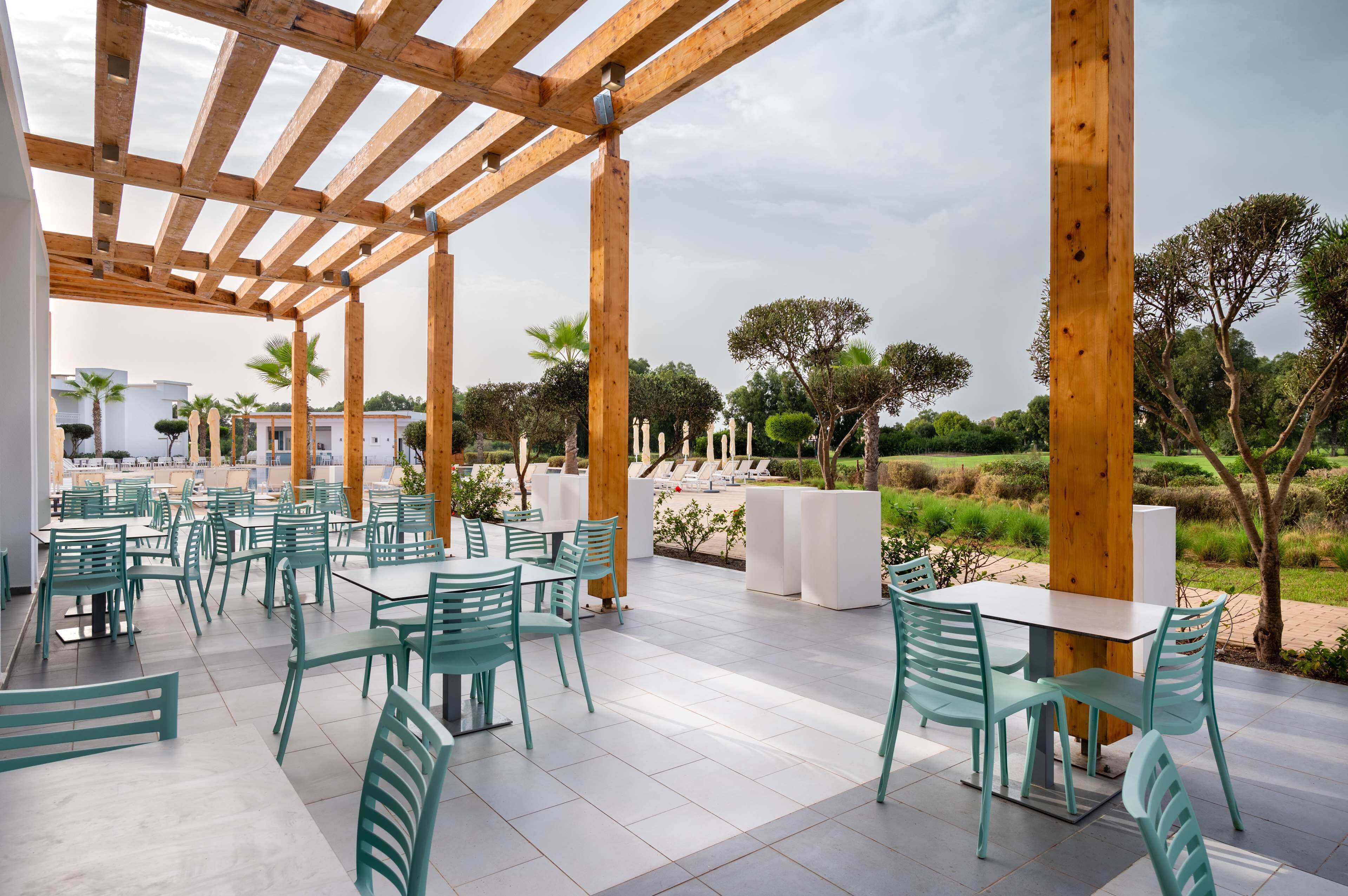 An outdoor patio with wooden pergola, green chairs, and white tables.
