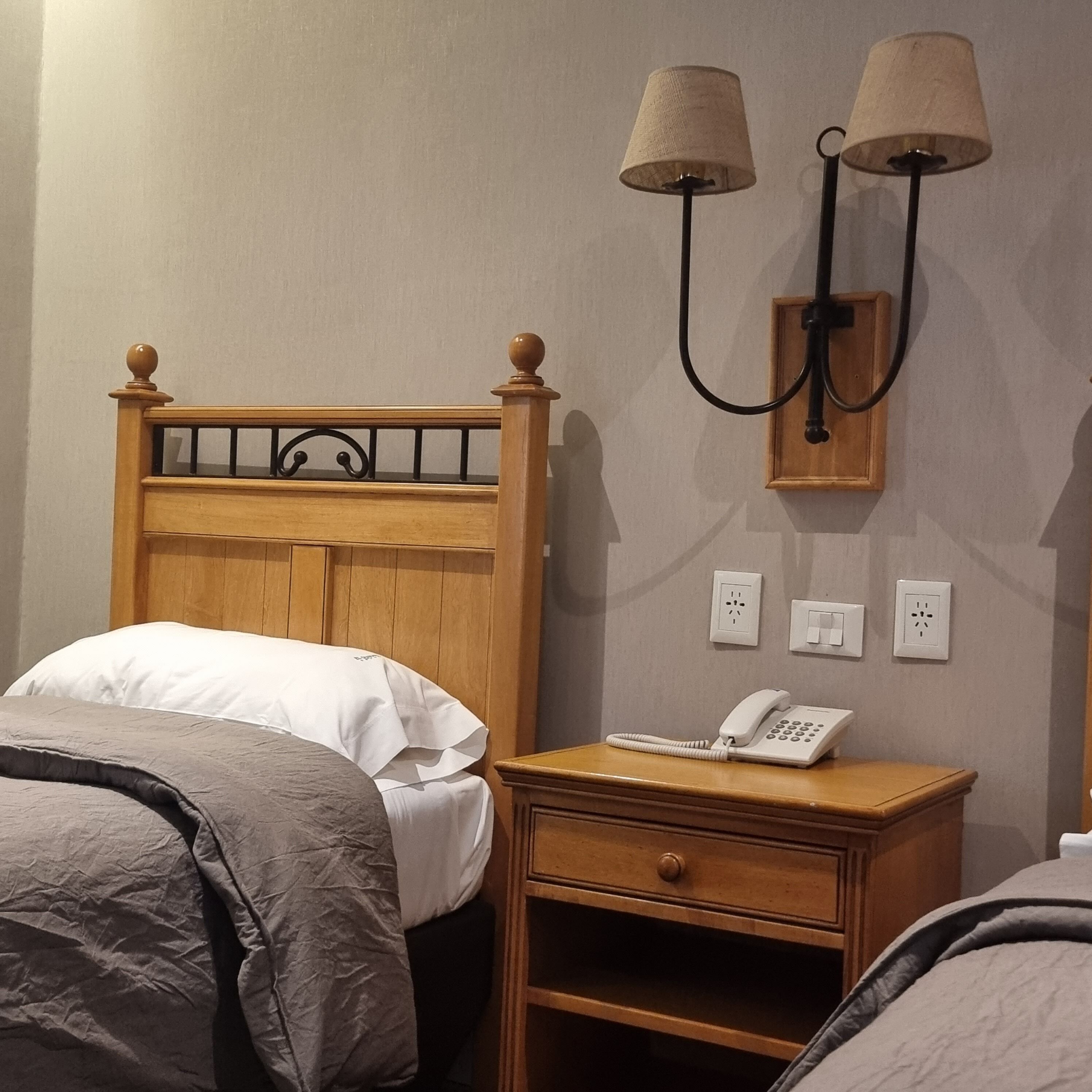 A neatly made bed with white and gray bedding, a wooden nightstand with a telephone, and two wall-mounted lamps.