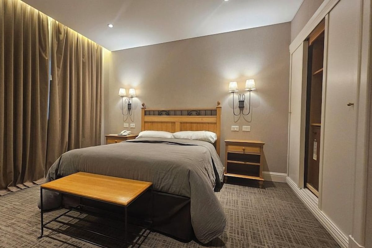 Ein Hotelzimmer mit Bett, Nachttischen, einer Bank, Vorhängen und Wandleuchten.
