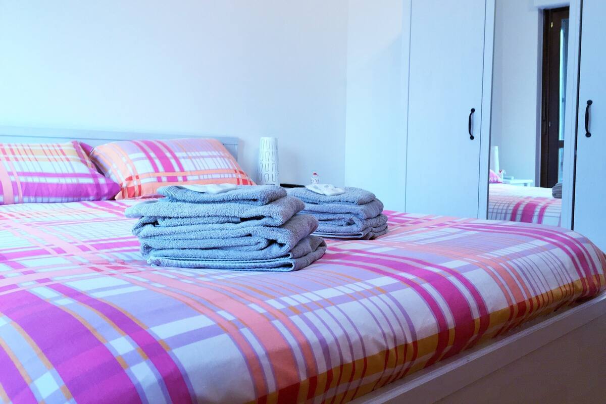 A neatly made bed with a colorful striped bedspread and pillows, a stack of folded towels, and a white bedside table with a lamp.