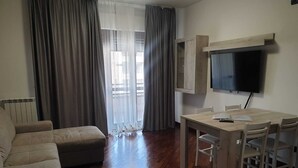 A living room with a sofa, a dining table with chairs, a TV mounted on the wall, and a window with curtains. - Domus Al Parco Guest House (Fiumicino)