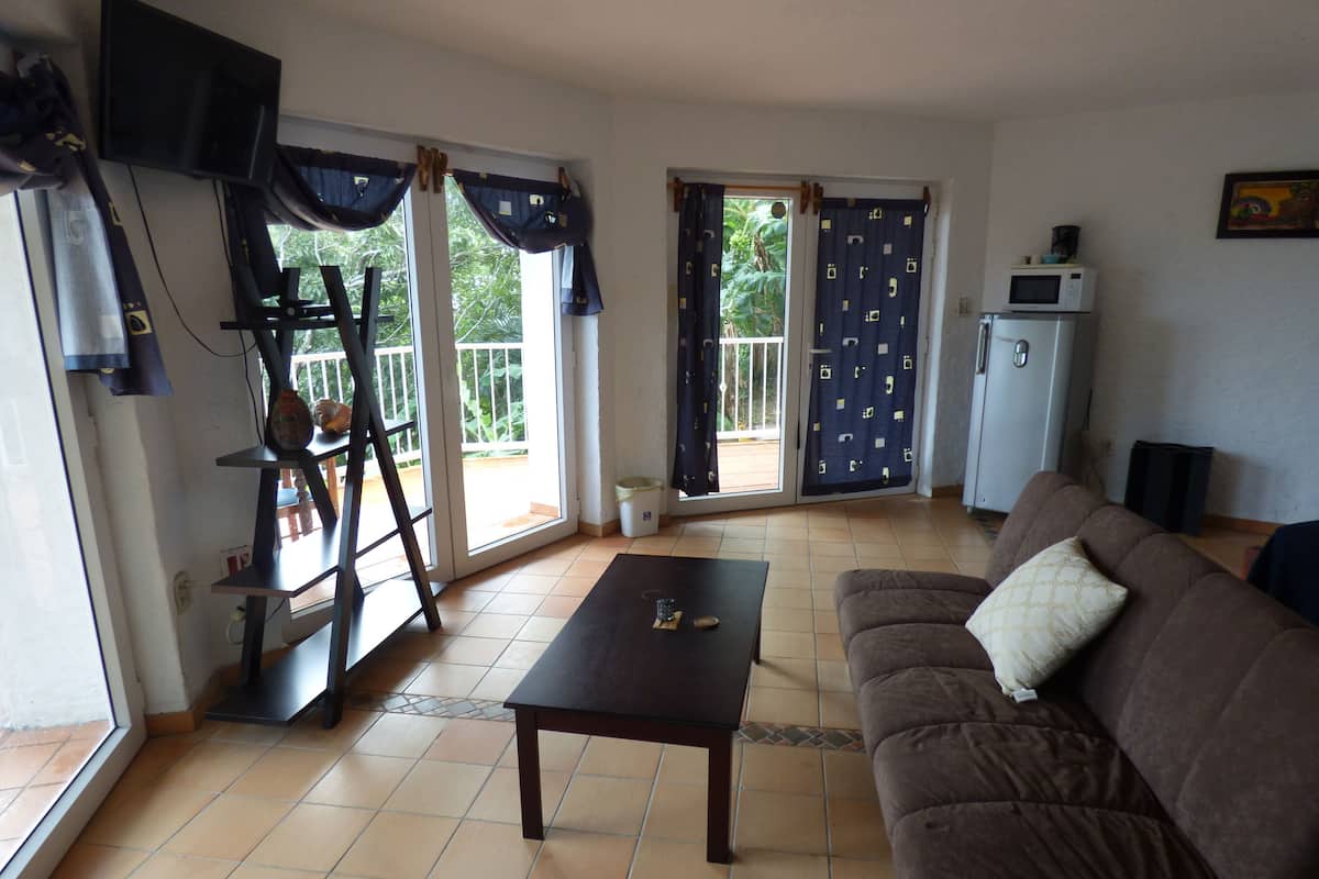 A living room with a brown sofa, a wooden coffee table, a TV mounted on the wall, and a sliding glass door leading to a balcony.