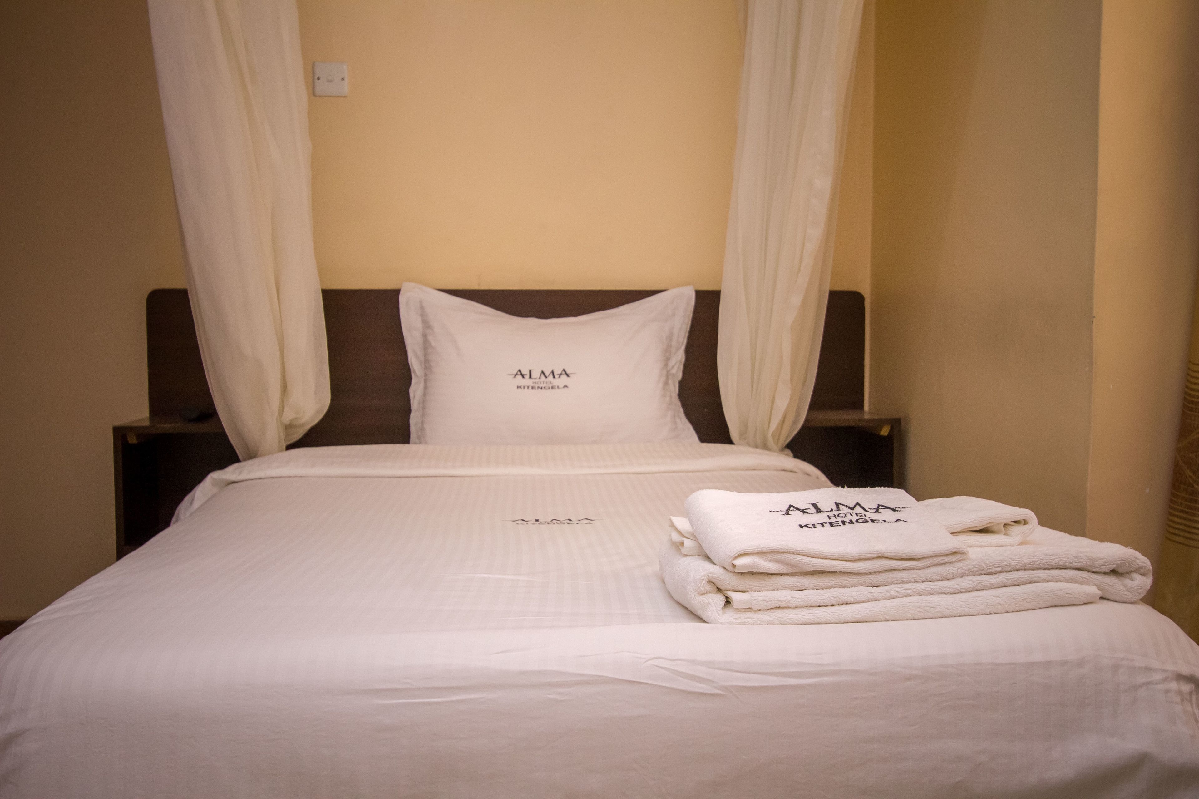 Un letto rifatto con cura, una coperta bianca, un cuscino con la scritta "ALMA HOTEL KITENGELA" e una pila di asciugamani bianchi.