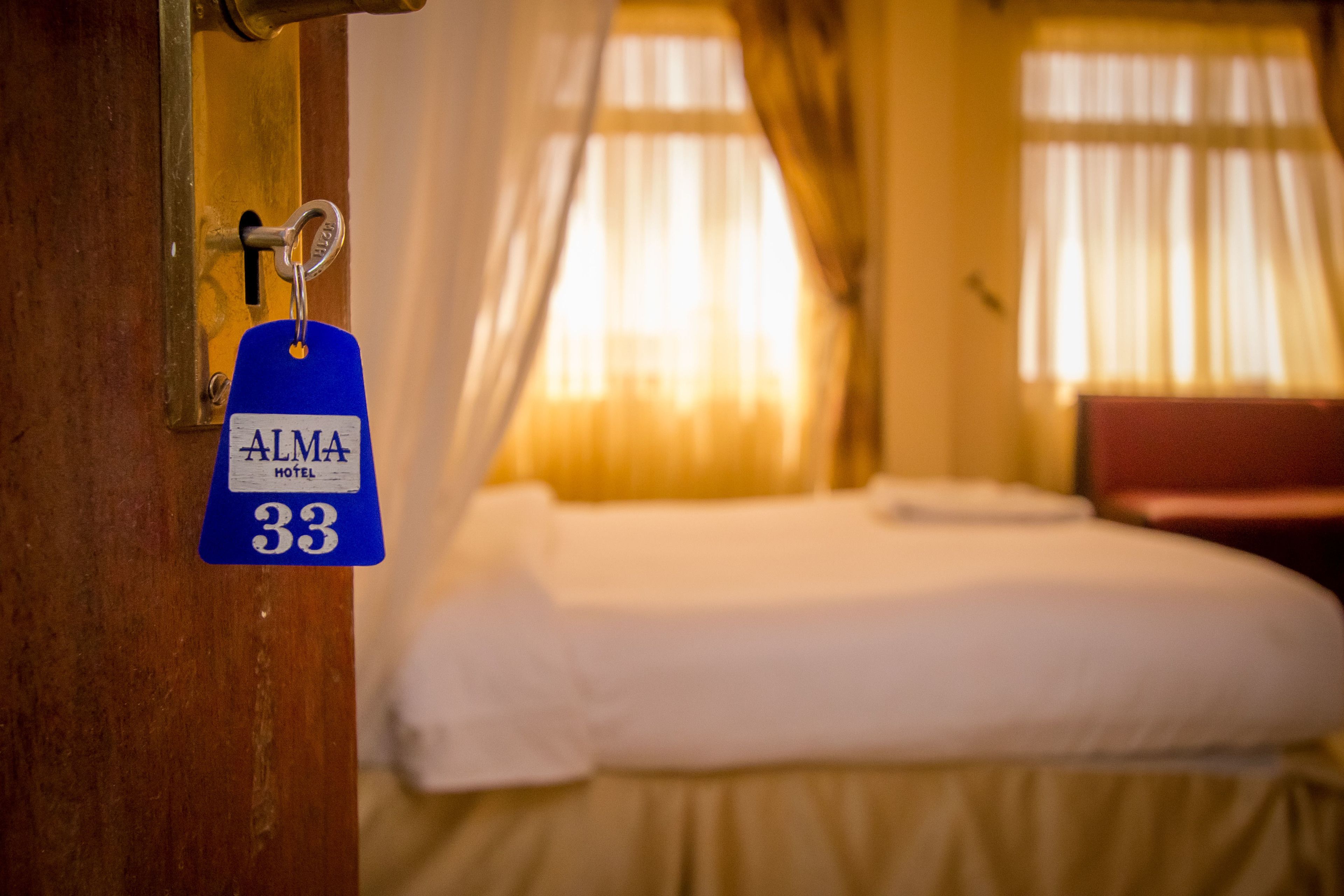 A hotel room with a bed, a key with a blue tag labeled "ALMA HOTEL 33", and a window with curtains.