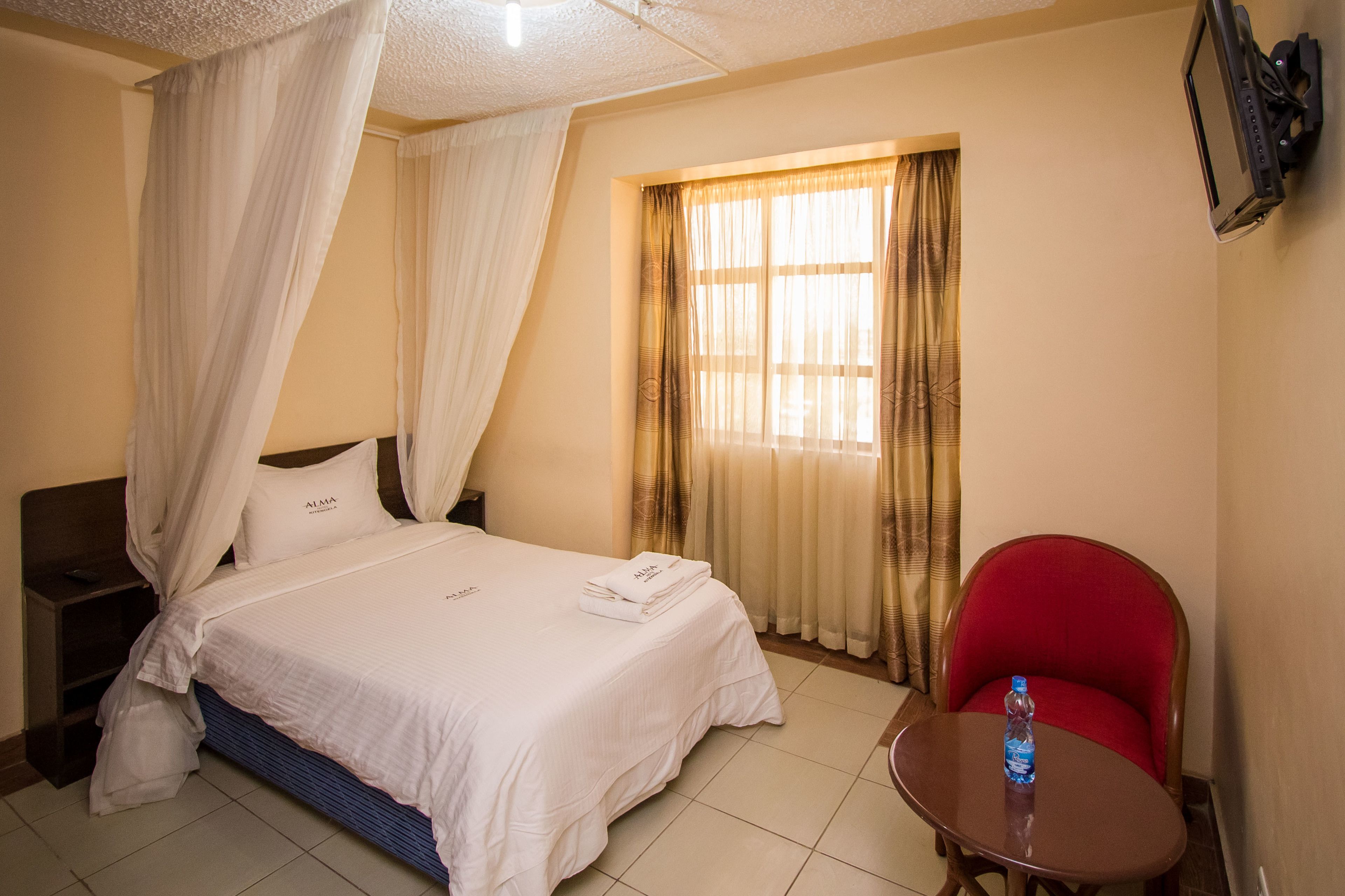 A hotel room with a bed, a nightstand, a red chair, a table with a bottle, and a TV mounted on the wall.