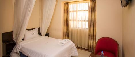 A hotel room with a bed, a nightstand, a red chair, a table with a bottle, and a TV mounted on the wall.