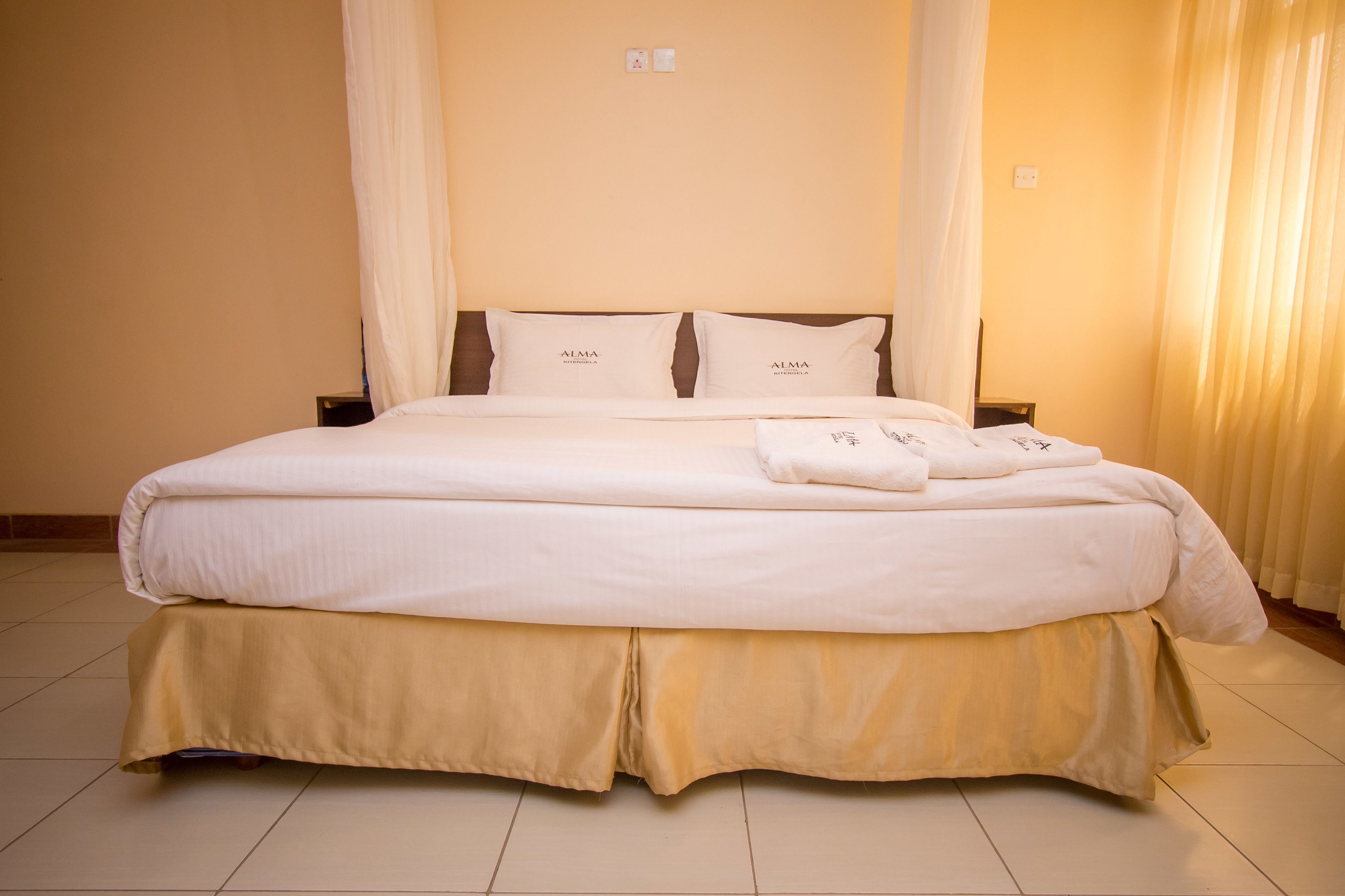 A neatly made bed with a beige bed skirt and pillows labeled "ALMA."