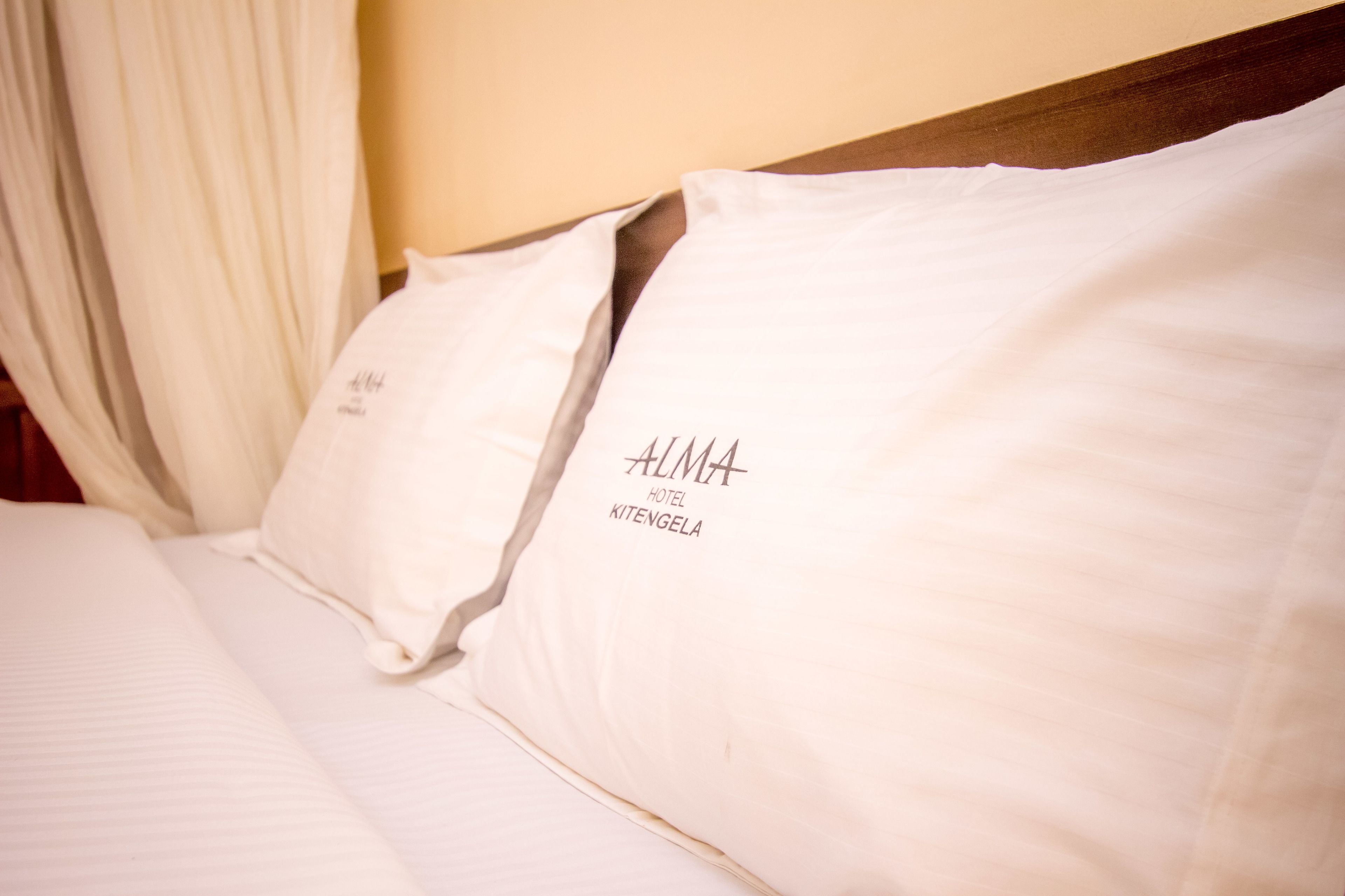 Ein Bett mit weißer Bettwäsche und Kissen, wobei eines das Schriftzug "ALMA HOTEL KITENGELA" trägt.
