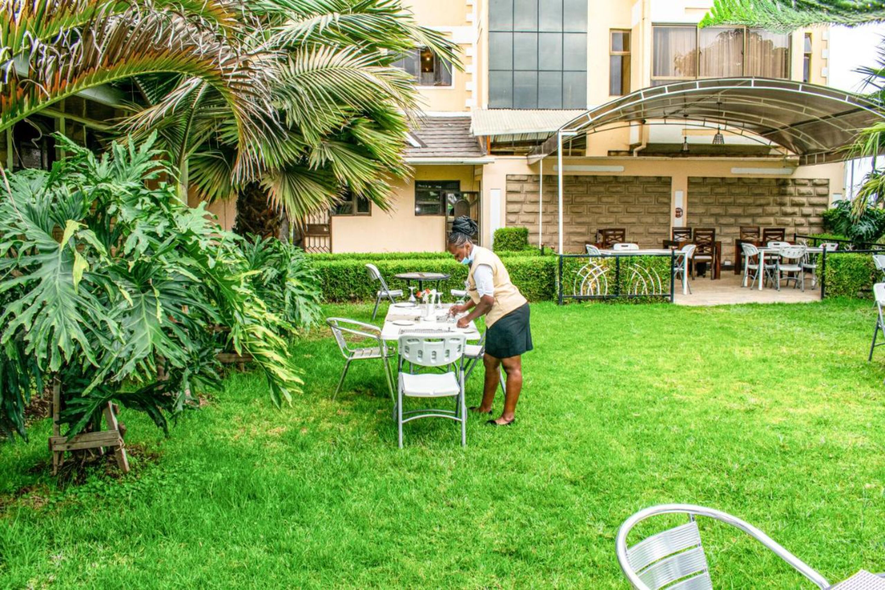 Uma área externa bem cuidada com espaço de estar coberto, uma mesa posta para refeições e vegetação exuberante, incluindo plantas de folhas grandes e uma palmeira.