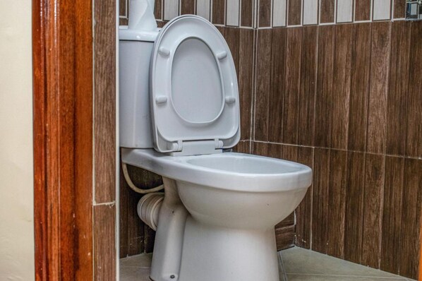 A white toilet with a closed seat in a bathroom with wooden panel walls. - Astorian Grand Hotel (Naivasha)