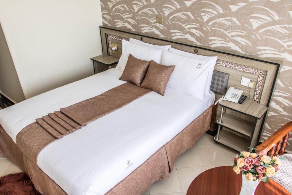 A neatly made bed with a brown bedspread and pillows, a wooden headboard, a nightstand with a phone and a small table, and a floral arrangement on a side table.