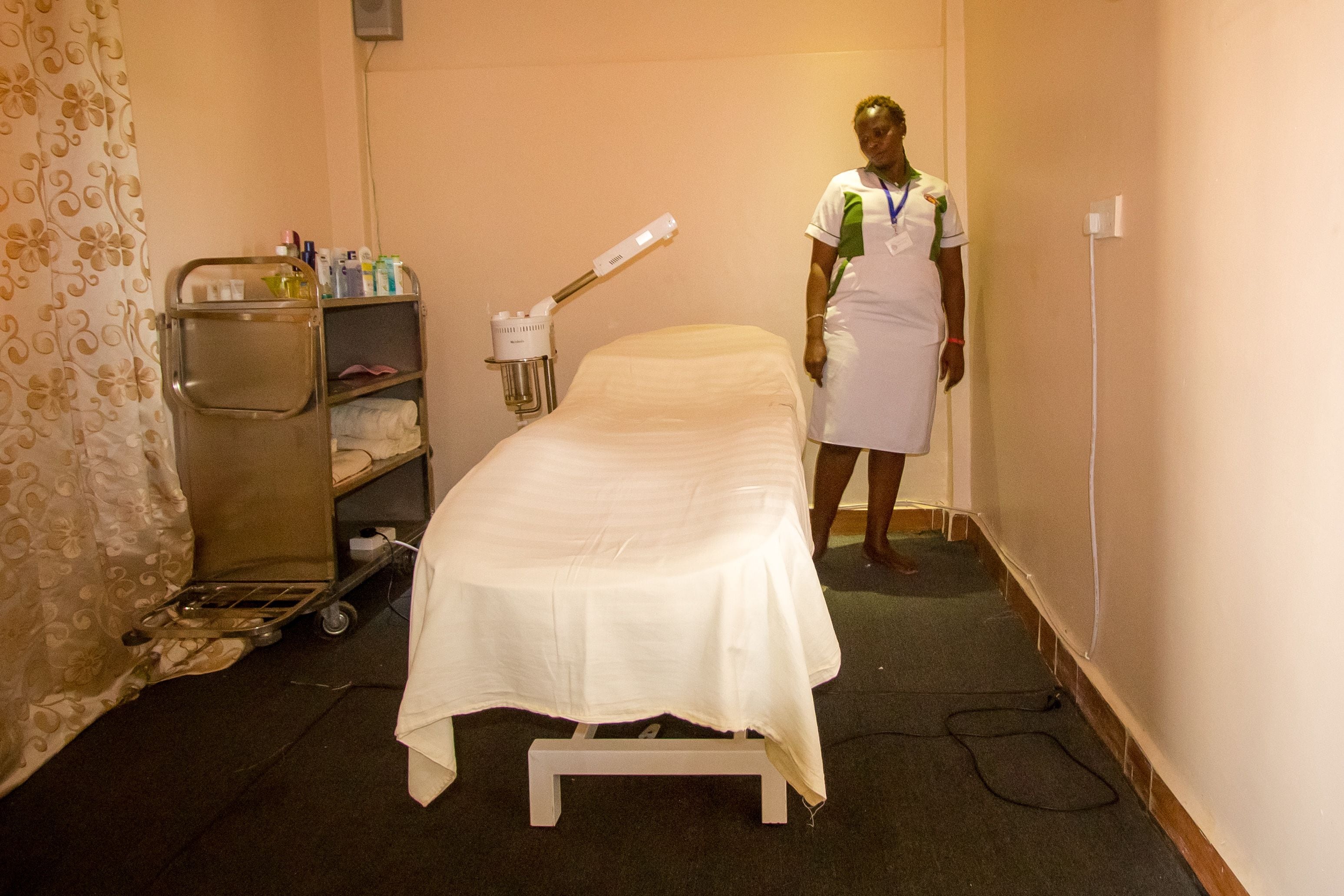 A small, narrow room with a single bed, a metal cart with toiletries, and a curtain with a floral pattern.