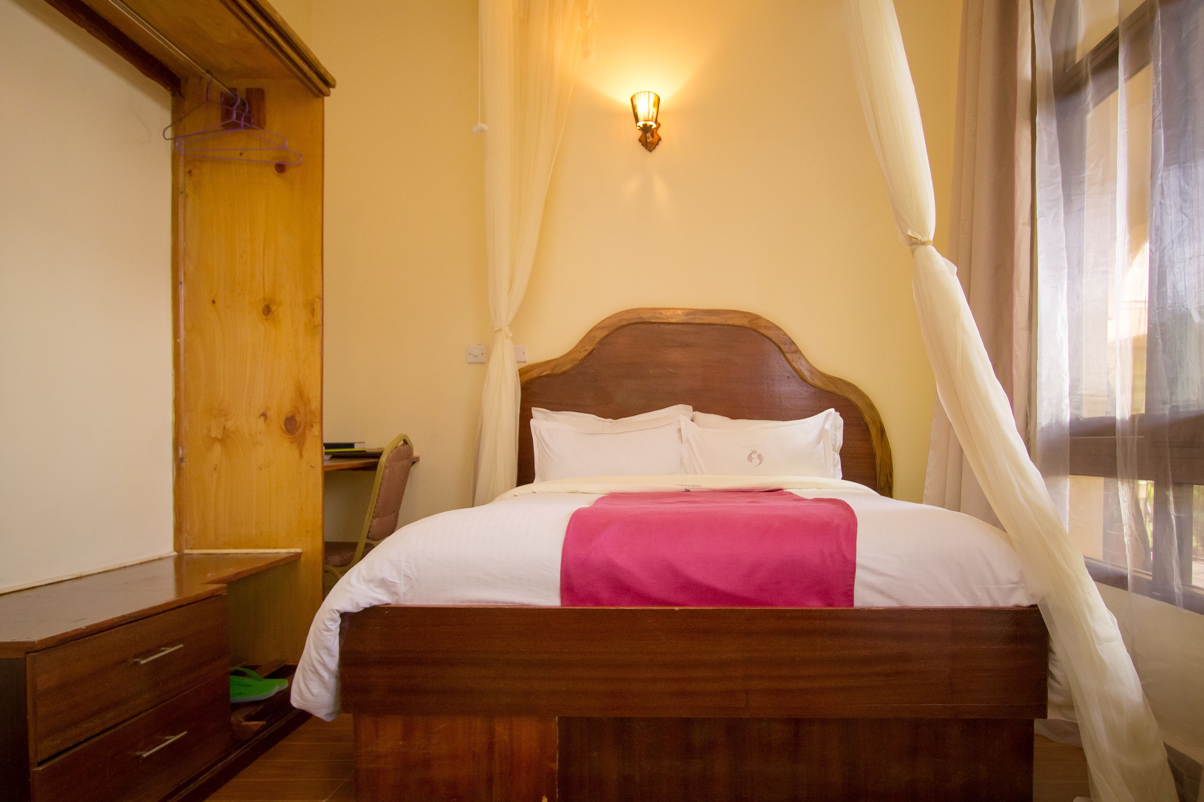 A wooden bed with white bedding and a pink blanket, a wooden wardrobe, a wall-mounted light fixture, and a window with sheer curtains.