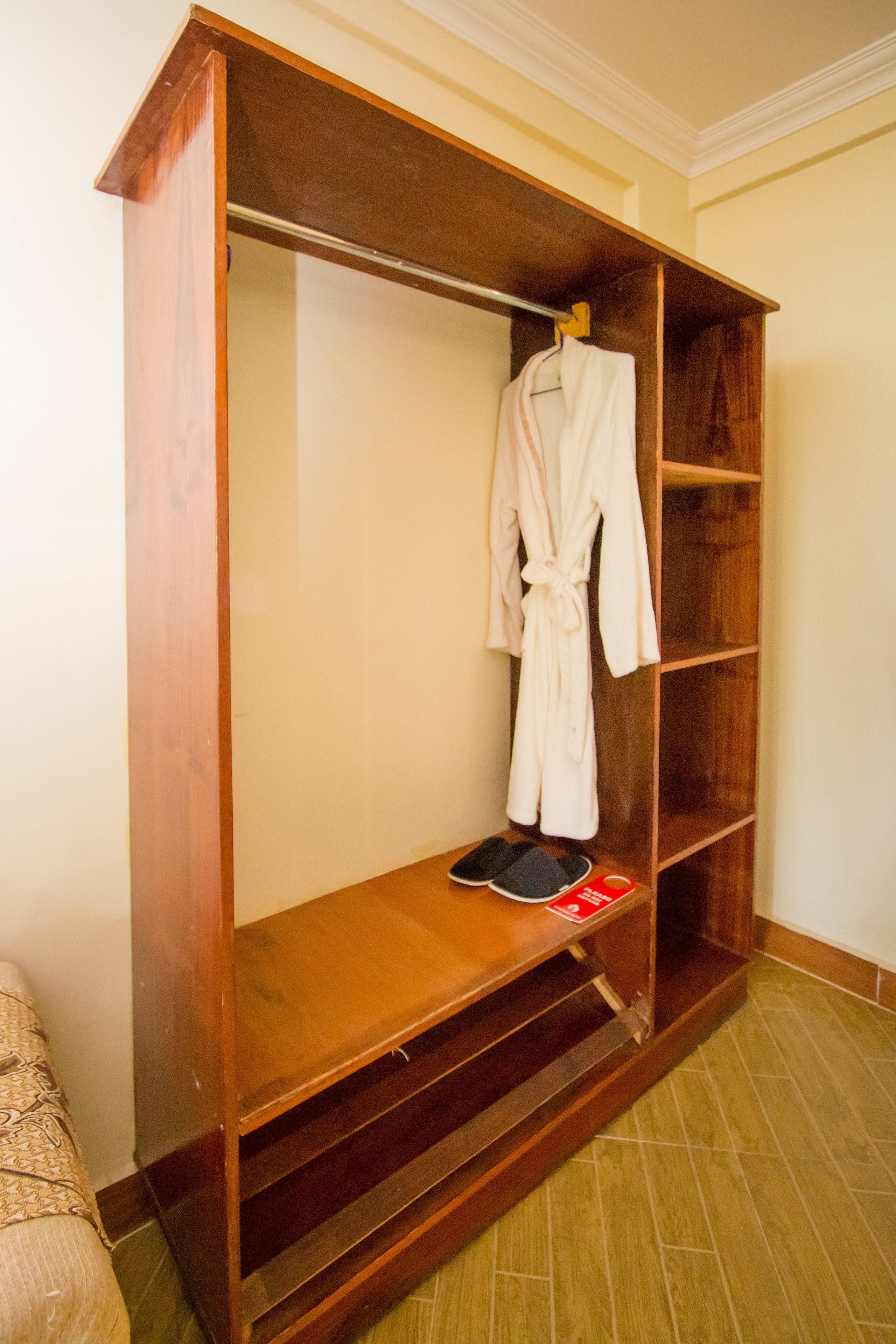 A wooden walk-in closet with a robe hanging on the door, a pair of slippers on the floor, and a shelf with a red object.