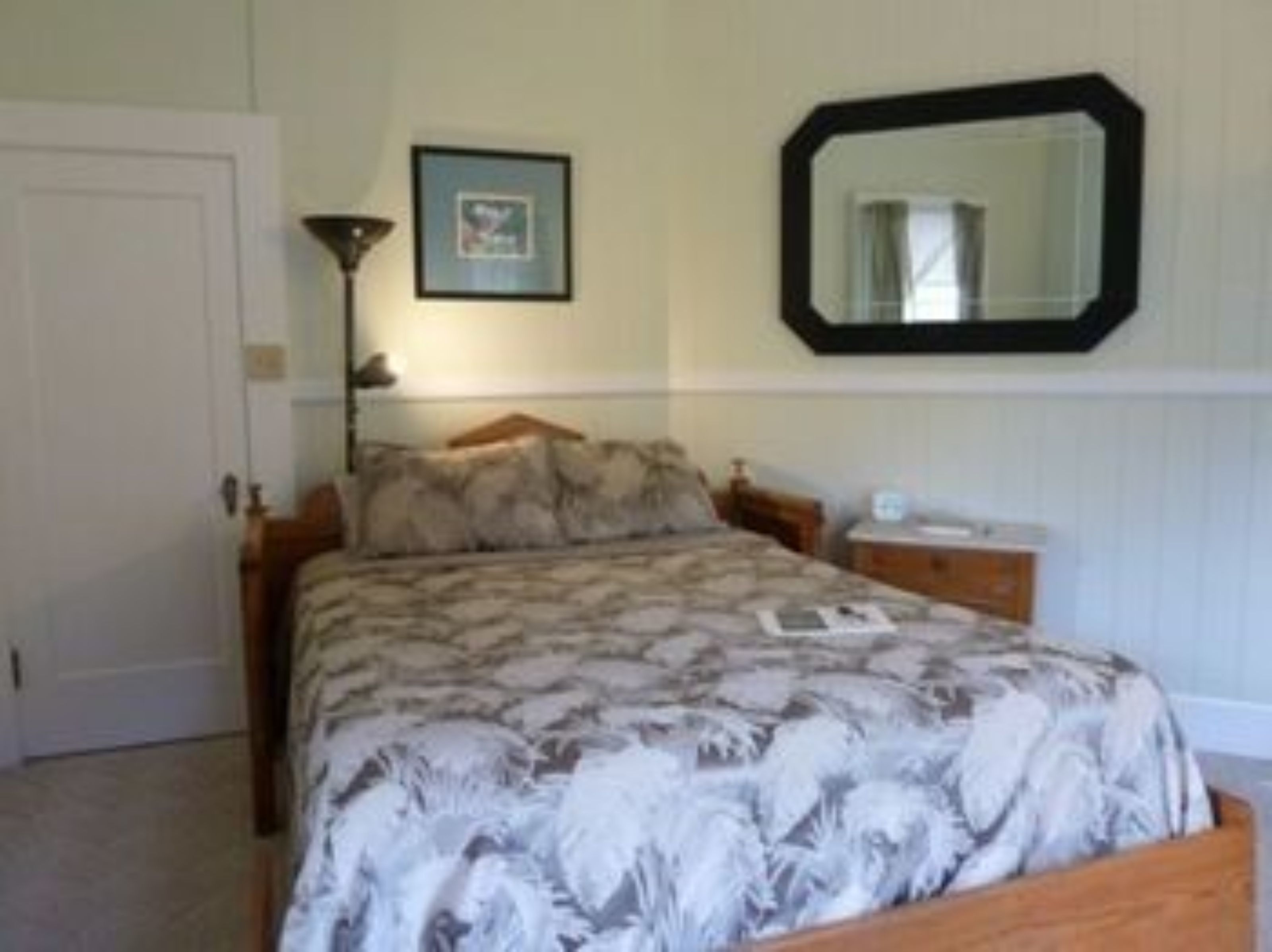 A bedroom with a bed, a bedside table, a lamp, a framed picture on the wall, and a mirror.