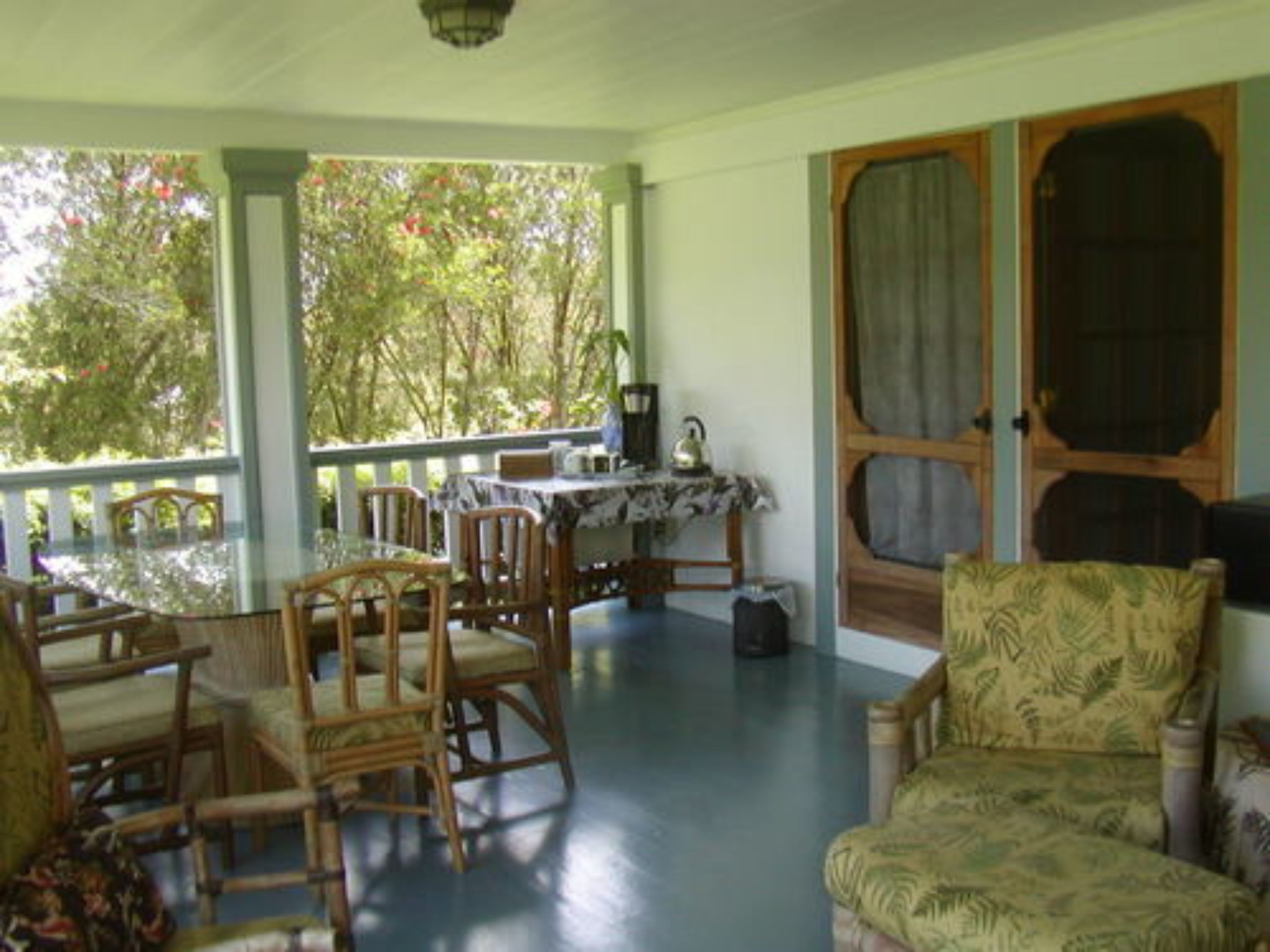 A covered porch with wooden chairs and a table, a door with a window, and a view of trees.