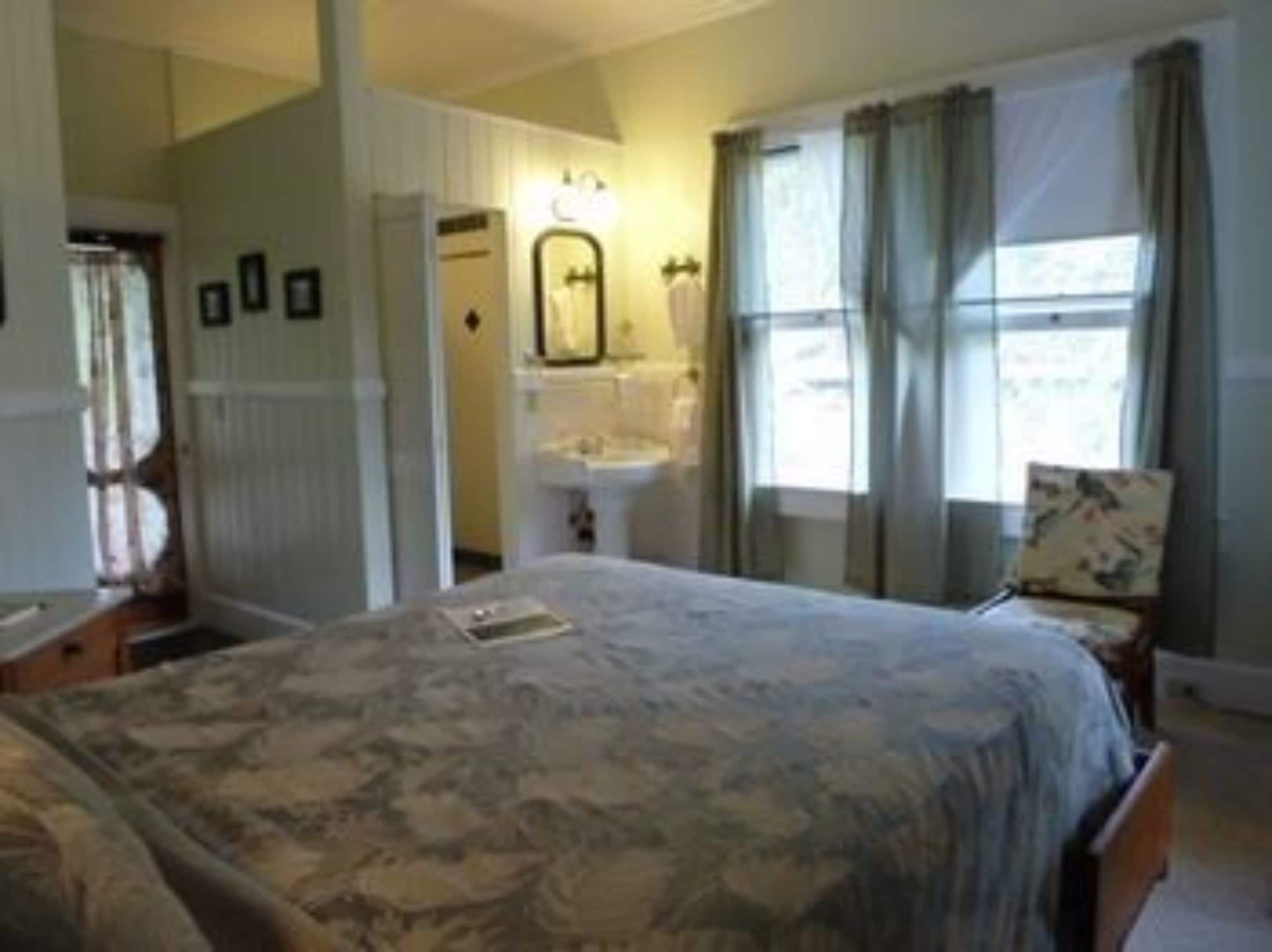 Une chambre à coucher comprenant un lit, une table de chevet, une chaise, une fenêtre avec des rideaux, un miroir et un luminaire fixé au mur.