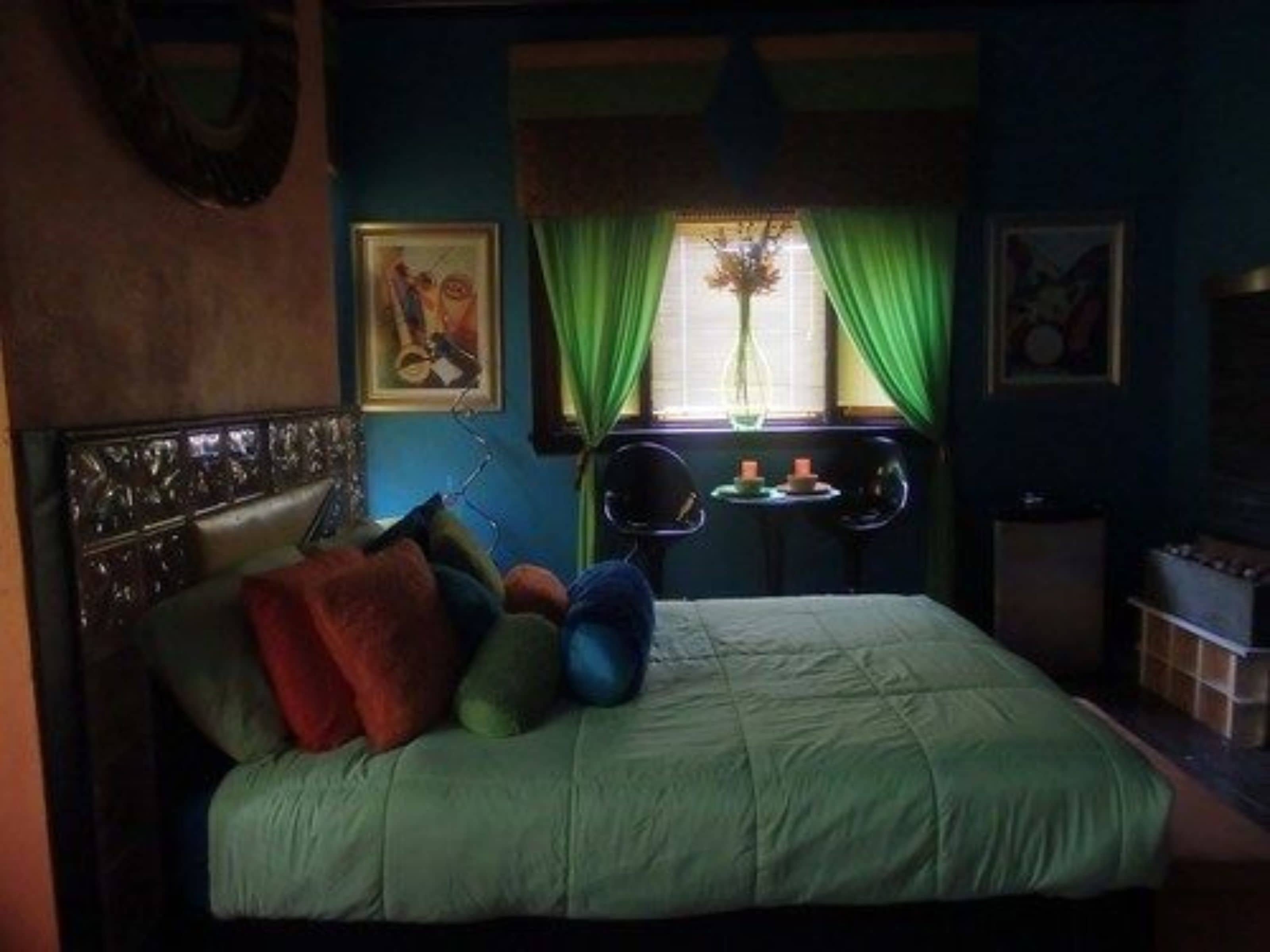 Un dormitorio con cama, cortinas verdes, una mesita con velas y un florero con flores.