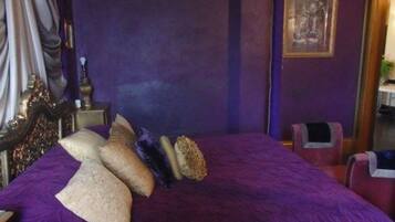 A room with a large bed, purple bedding, a nightstand, and a framed picture on the wall.