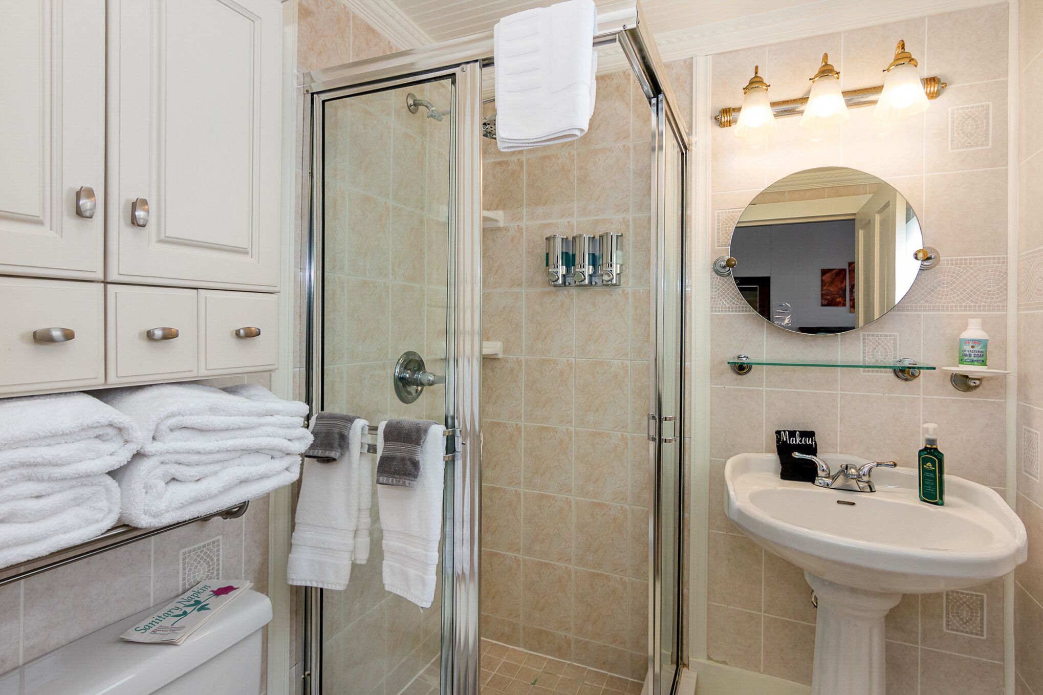 A bathroom with a glass shower enclosure, a sink with a mirror, and neatly folded towels on a shelf.