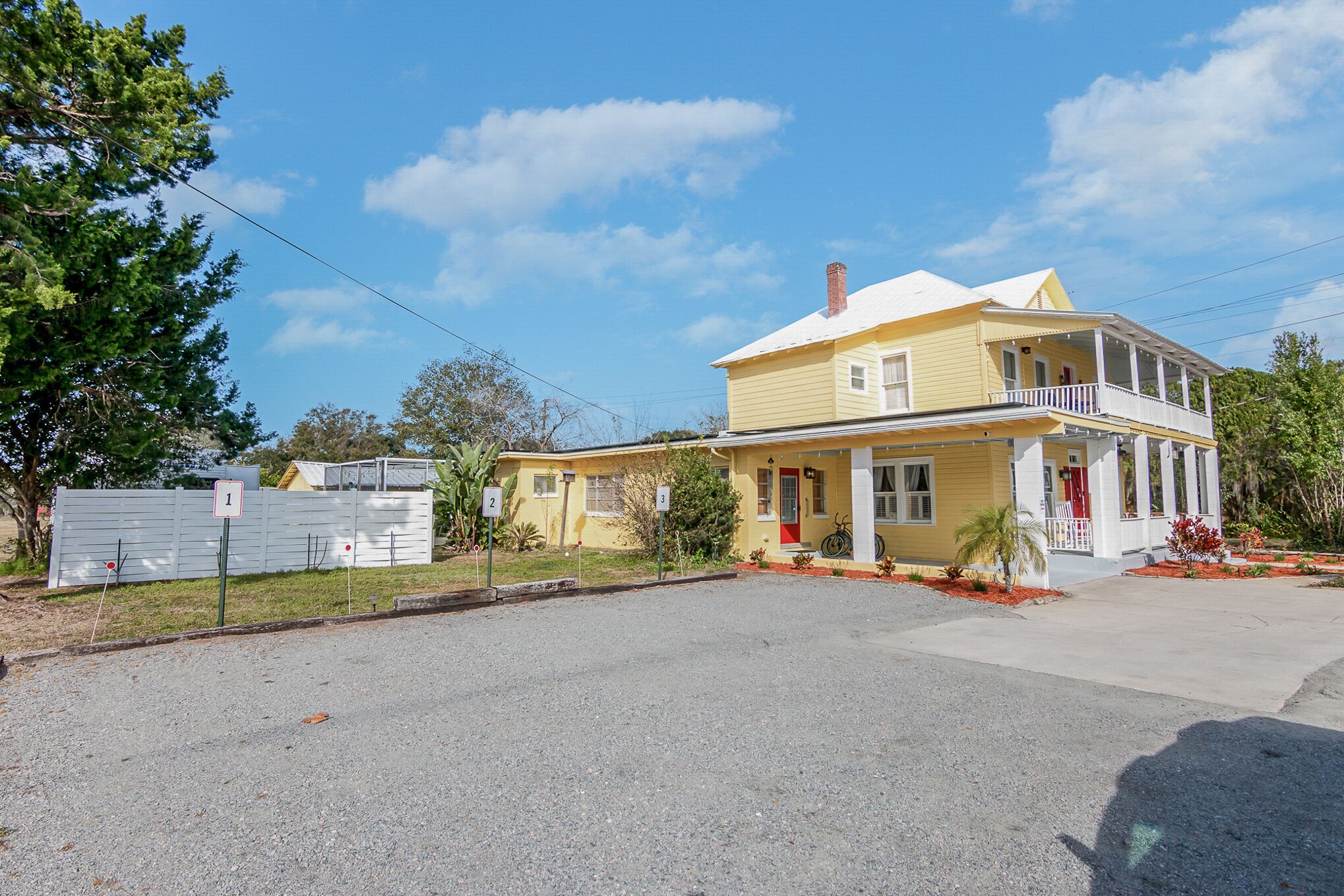 Uma casa amarela de dois andares com varanda, quintal cercado e entrada de cascalho.