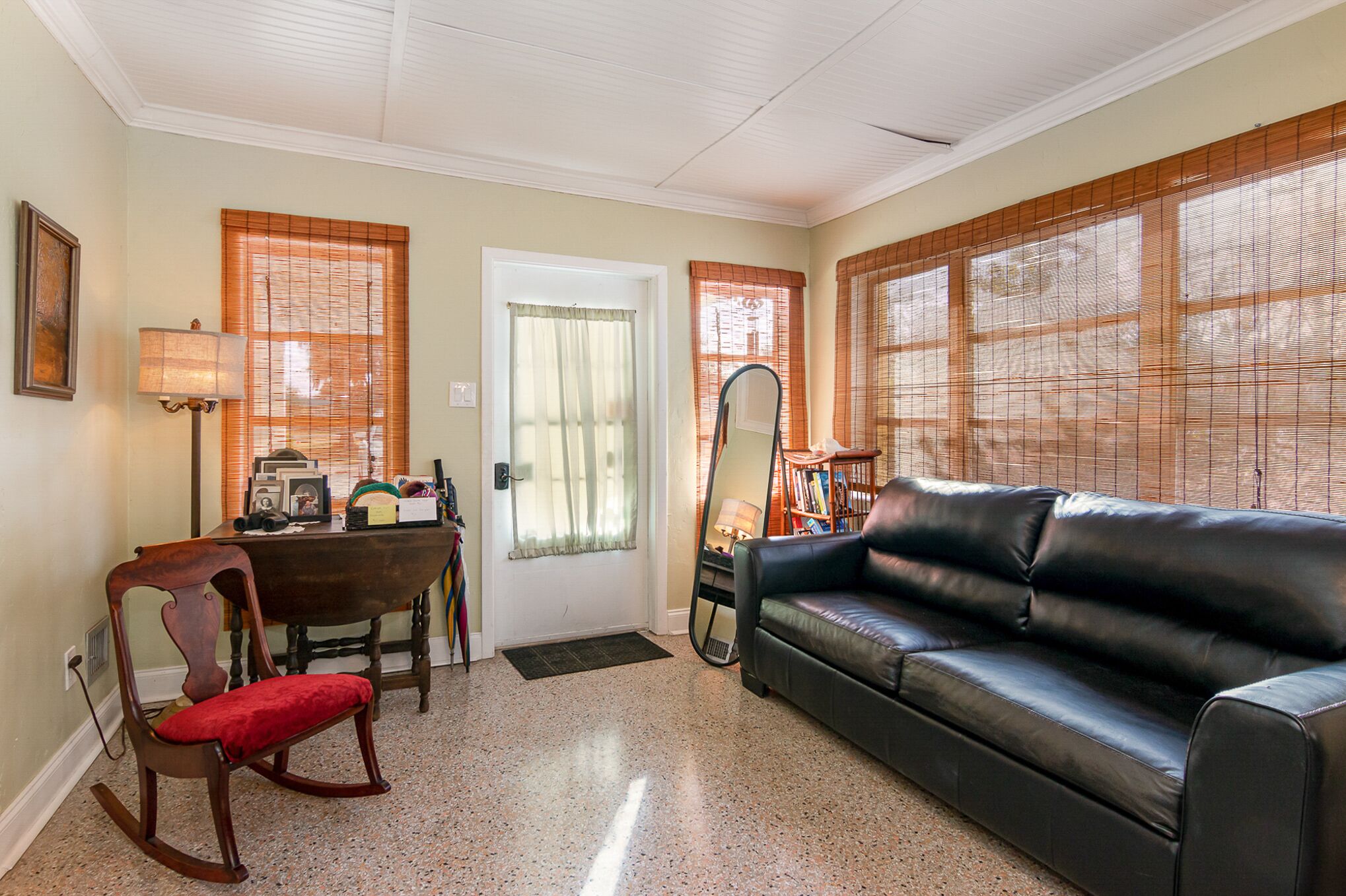 A living room with a black leather sofa, a wooden rocking chair, a dining table with chairs, and a large mirror.