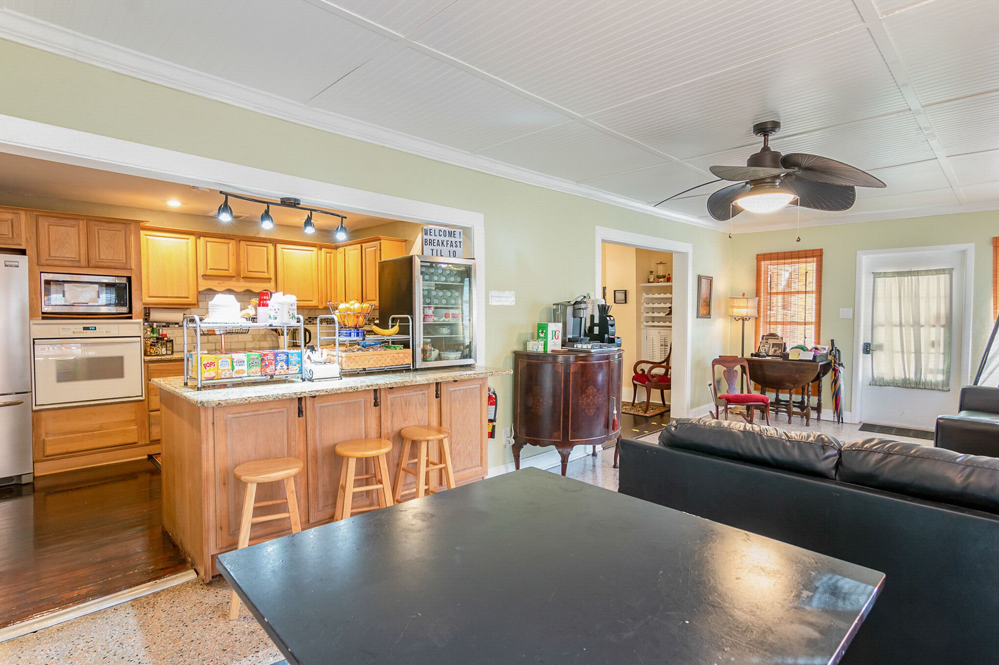 A kitchen with wooden cabinets, a microwave, and a breakfast bar with stools. There is a ceiling fan and a dining area with a table and chairs.