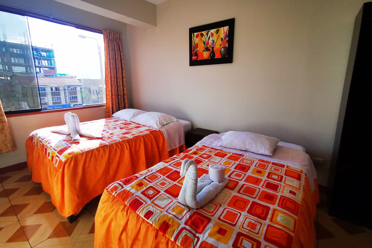 Quarto de hotel com duas camas, cada uma com colcha laranja e almofadas brancas, e uma janela com vista para prédios.