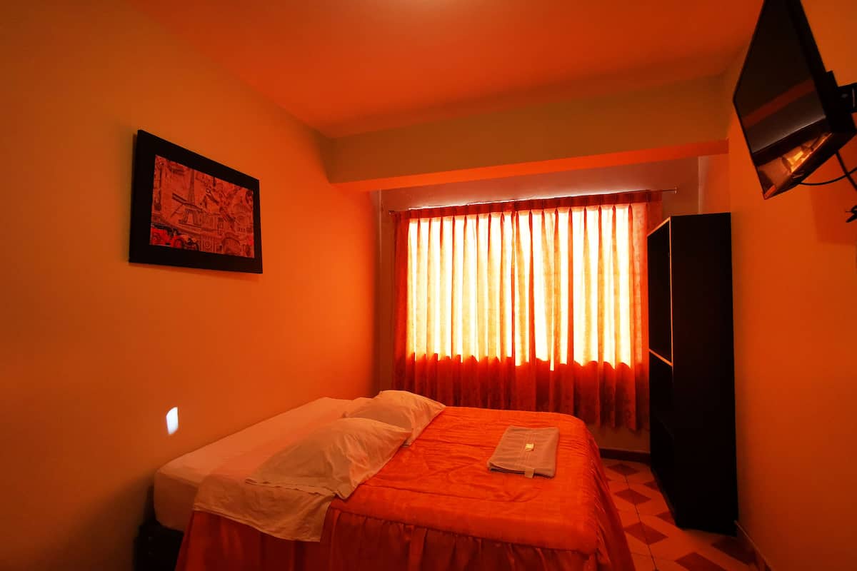 Um quarto pequeno e com pouca luz, com uma cama de solteiro, um quadro na parede e uma televisão fixada na parede.