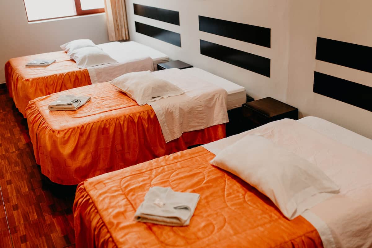 Quarto de hotel com três camas de solteiro, cada uma com colcha laranja e travesseiros brancos. As camas estão dispostas em linha, encostadas a uma parede com cabeceiras pretas.