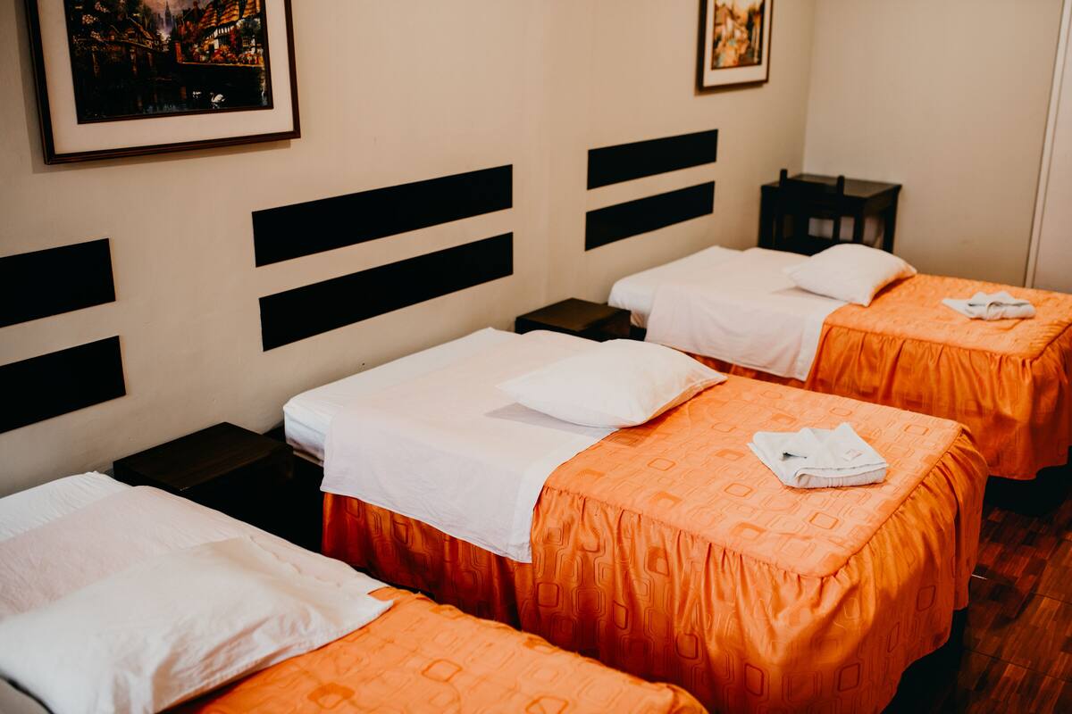 Quarto de hotel com três camas, cada uma com almofada branca e colcha laranja. Há cabeceiras pretas, uma pequena mesa de cabeceira e uma moldura com um quadro na parede.