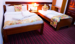 A hotel room with two wooden beds, each with a floral bedspread and pillows. There is a nightstand with a lamp, a telephone, and a book. The room has a red striped carpet and wooden furniture. - Wida Highway Motel (Kikuyu)