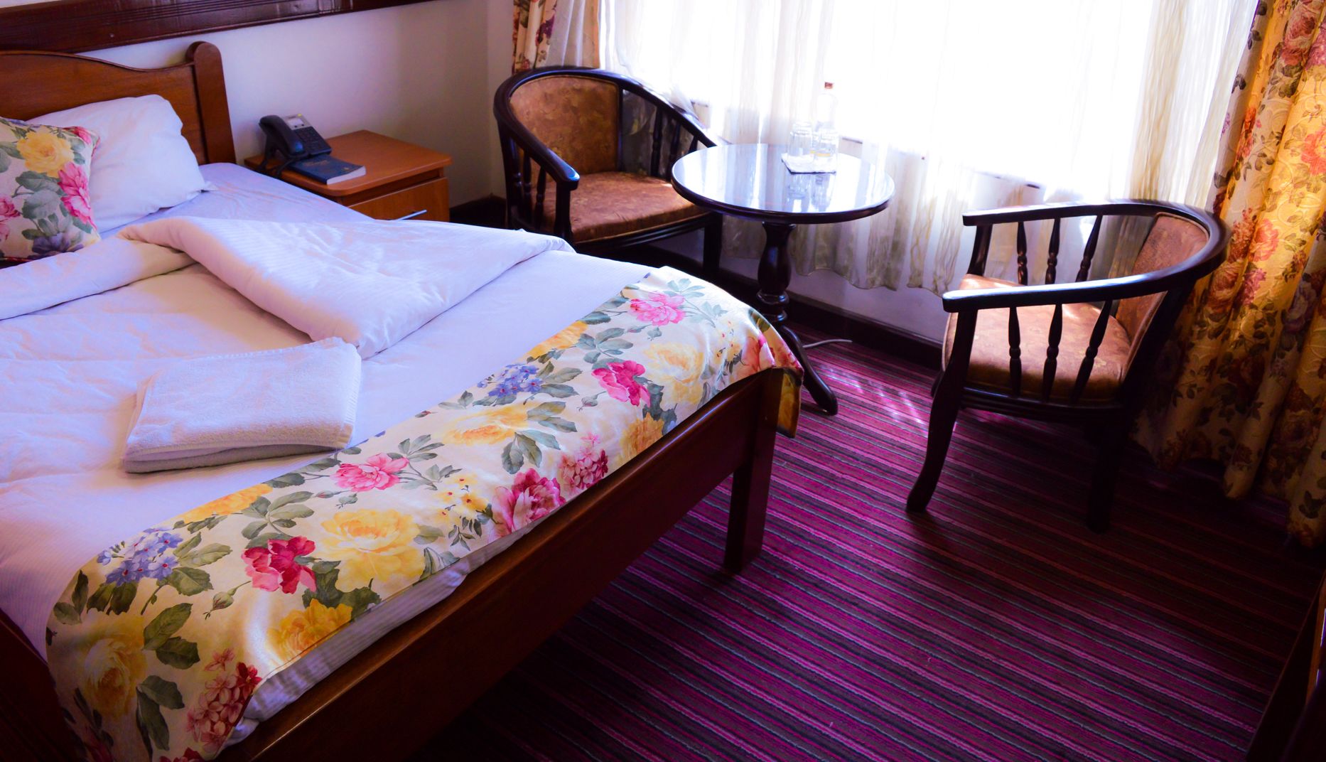 Une chambre d  ’hôtel avec un lit, une table de chevet avec un téléphone, une chaise, une petite table avec un verre et une fenêtre avec des rideaux.