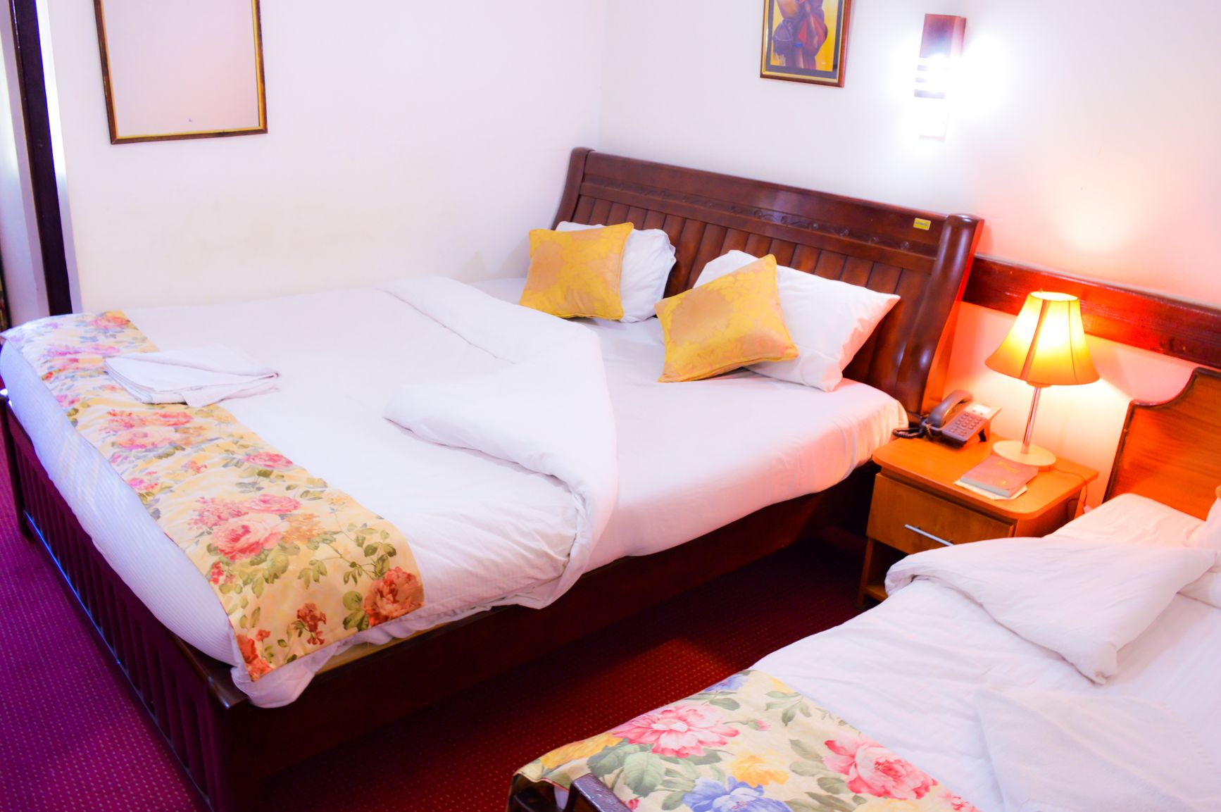 A hotel room with two beds, each with floral bedding and pillows, a wooden headboard, bedside tables with lamps, and a red carpet.