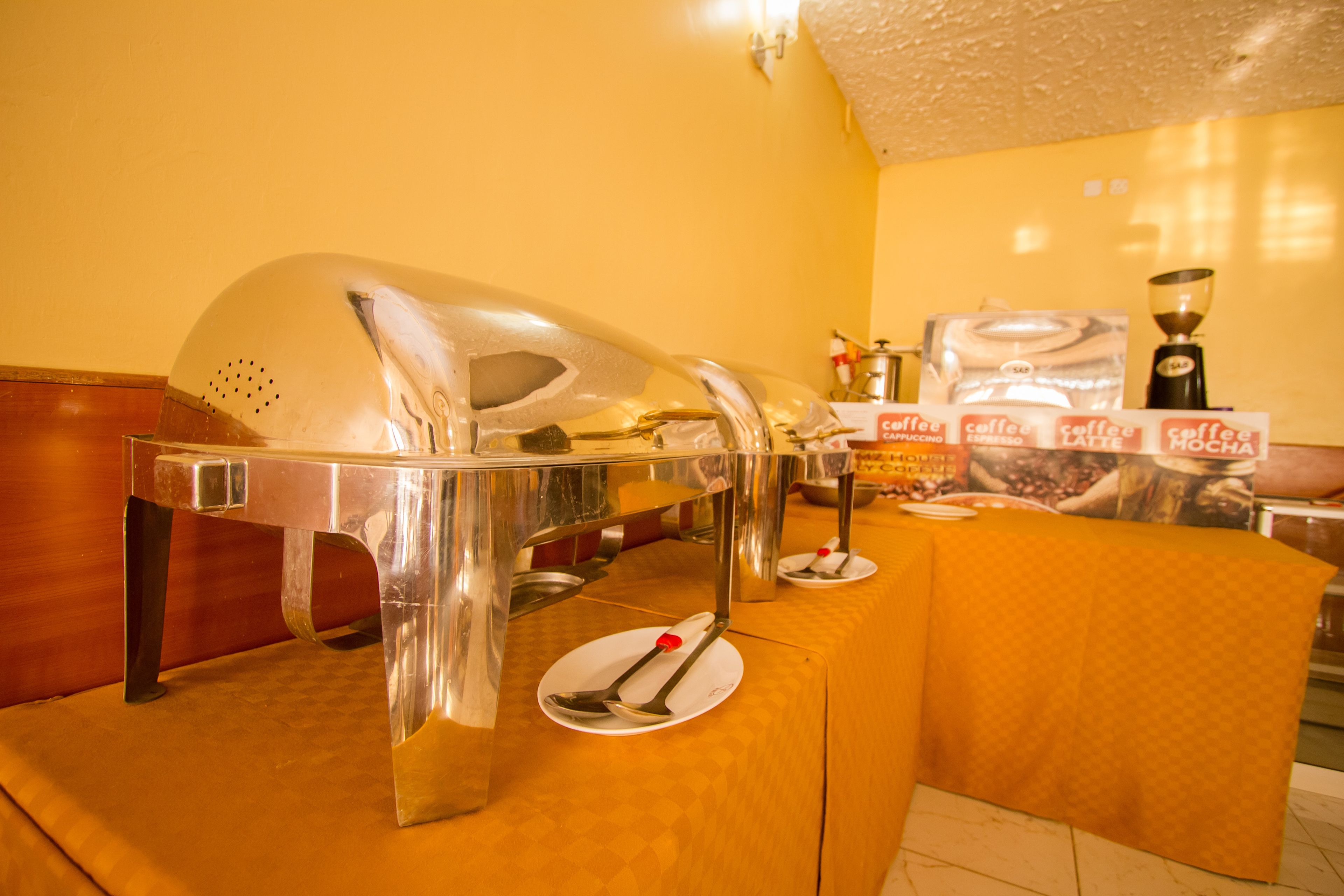 A buffet setup with a clear glass serving station, a coffee station with branded packaging, and a table with a yellow tablecloth.