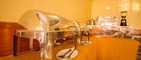 A buffet setup with a clear glass serving station, a coffee station with branded packaging, and a table with a yellow tablecloth.