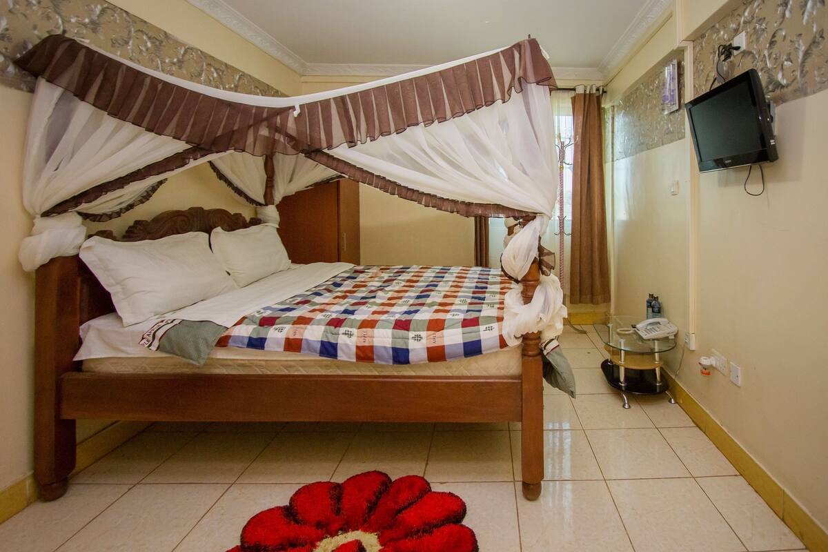 A bedroom with a bed, a TV mounted on the wall, and a small table with a phone and a bottle.
