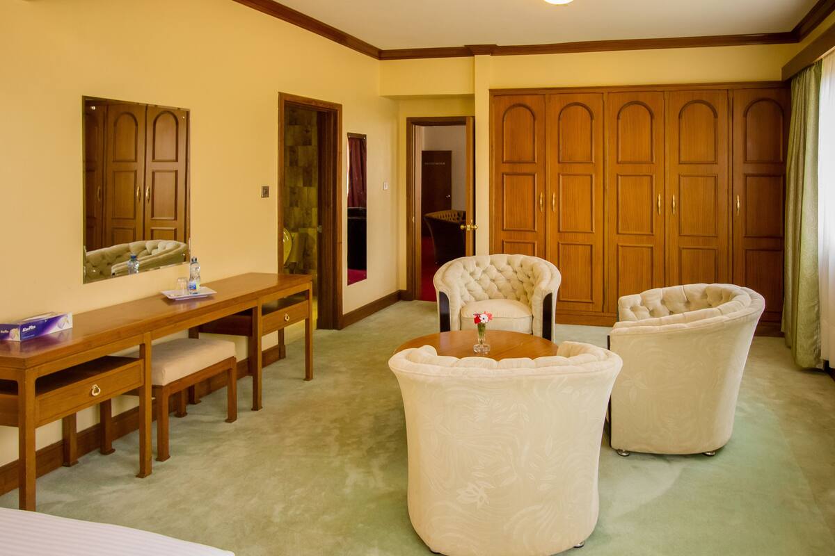 A hotel room with a wooden desk, two armchairs, a large wardrobe, and a mirror on the wall.