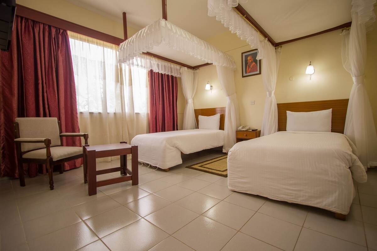 A hotel room with two beds, a wooden chair, a nightstand, and a wall-mounted lamp.