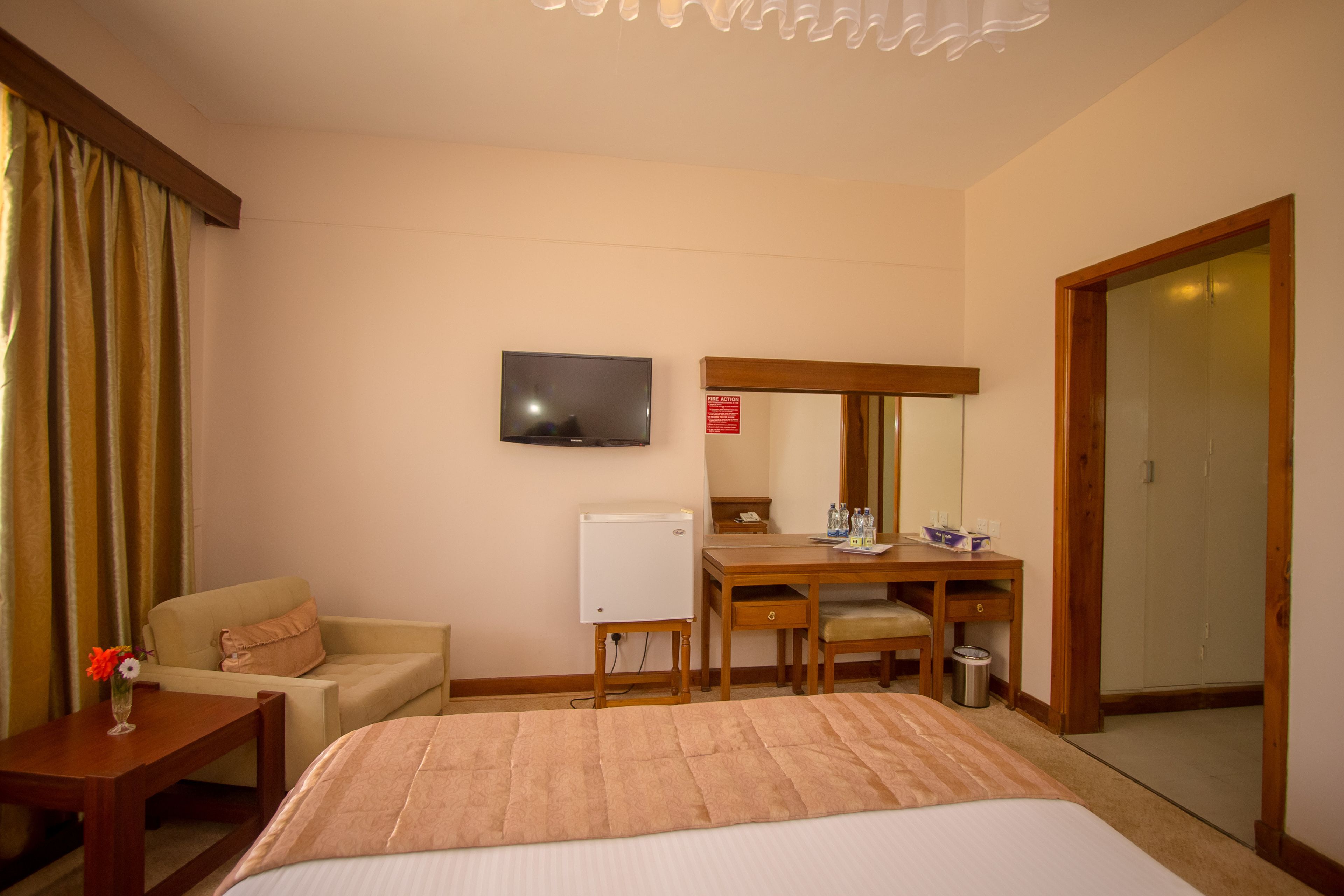 Una camera d'albergo con un letto, una scrivania, una sedia, una televisione, uno specchio, un armadio e un tavolino con un vaso di fiori.