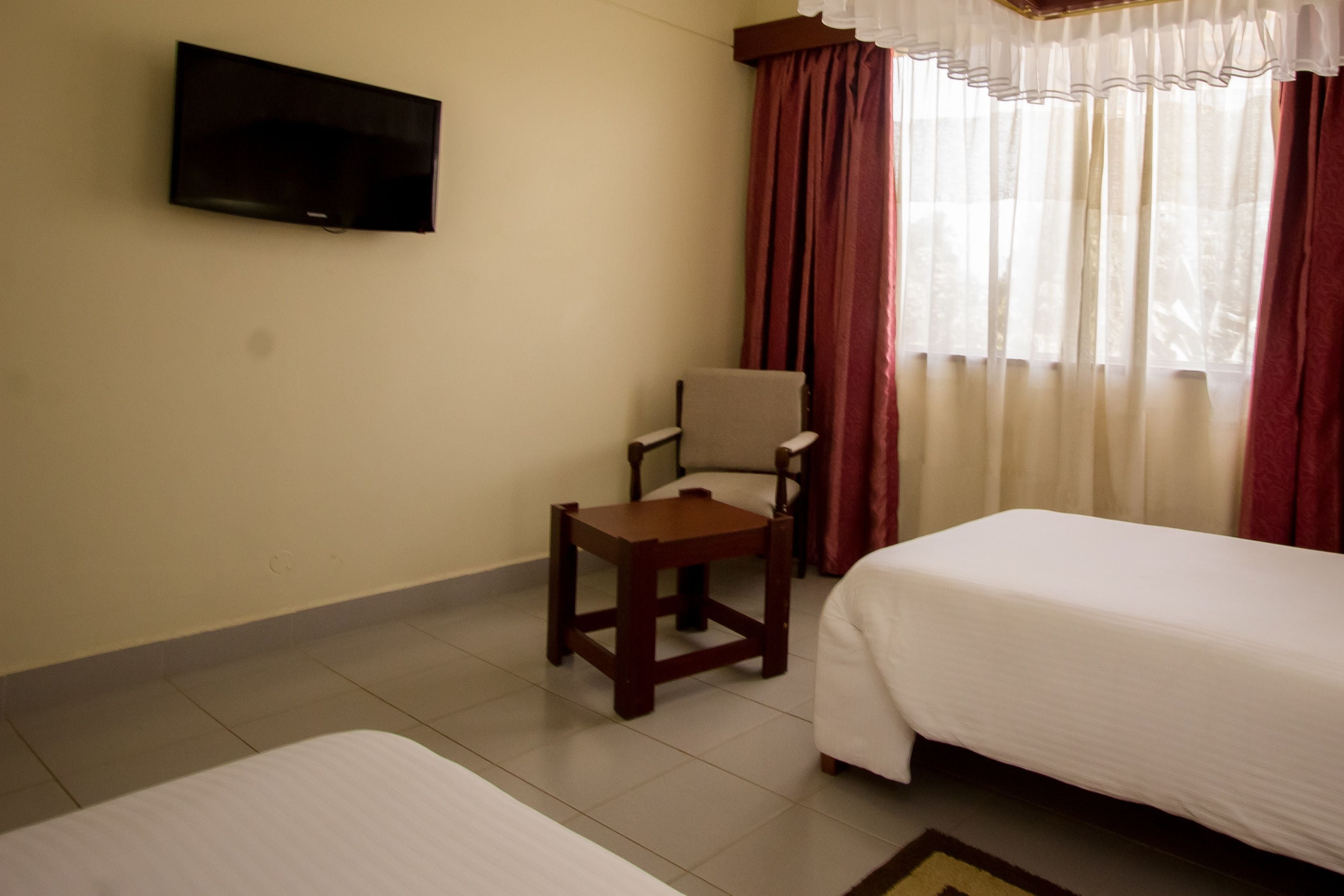 Quarto de hotel com cama, uma cadeira, uma mesinha, televisão fixada na parede e janela com cortinas.