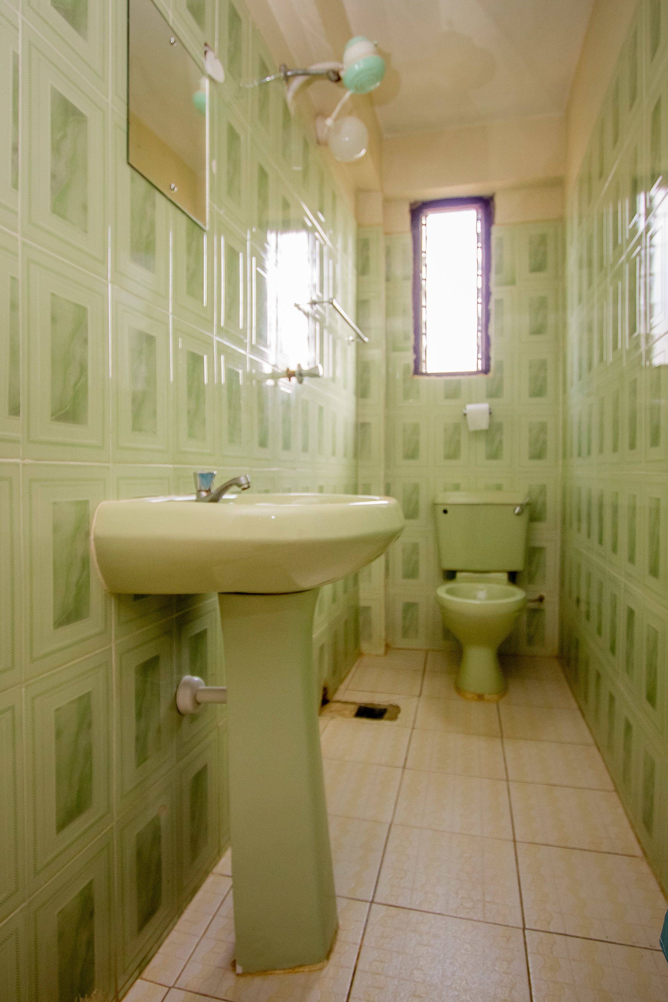 Un baño con paredes de color verde con diseño, un lavamanos, un inodoro y una ventana con marco morado.