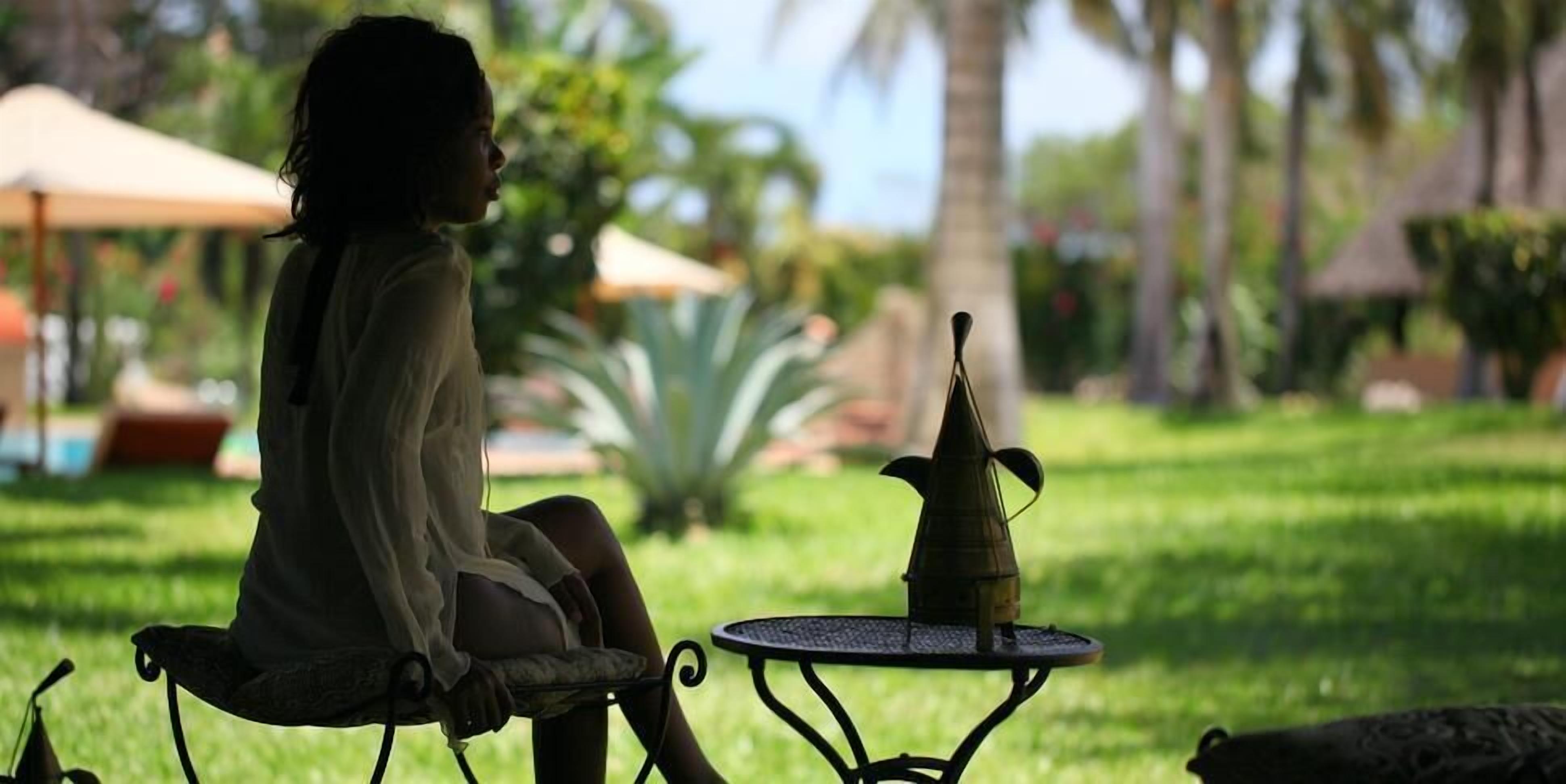 A person sitting on a chair at a table with a decorative object on it, in a garden with grass and palm trees.