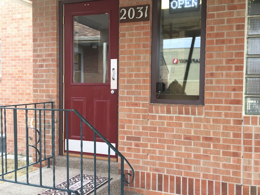 A brick building with a red door, a small window with an 'OPEN' sign, and a number '2031' on the wall.
