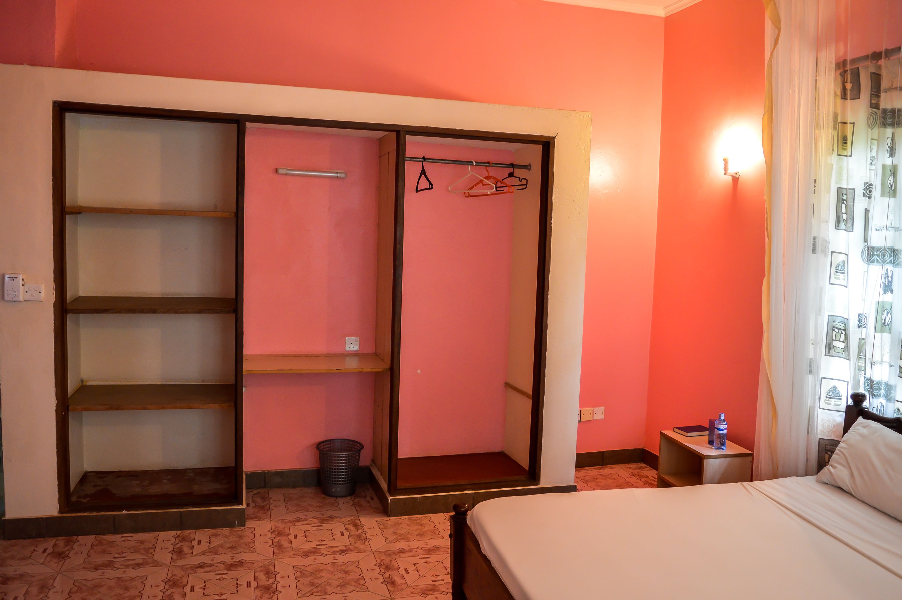 Hotelkamer met een bed, een garderobe met planken, een klein tafeltje met een waterfles en een wandlamp.