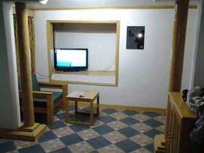 Una habitación pequeña con una televisión sobre una estantería, una silla, una mesita y un suelo estampado.