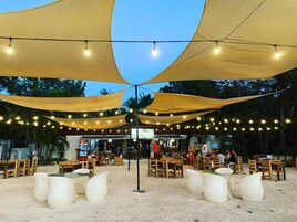 Un área de estar al aire libre con mesas y sillas bajo toldos amarillos, luces colgantes y vistas a la playa y palmeras.