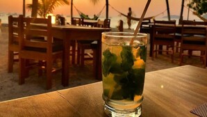 Un verre d’une boisson glacée, garni de feuilles de menthe, est posé sur une table en bois dans un restaurant en bord de mer, avec des chaises en bois et une vue sur le coucher de soleil.