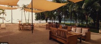 Un área de estar al aire libre con mobiliario de madera y toldos amarillos, rodeada de palmeras y vistas a la playa.